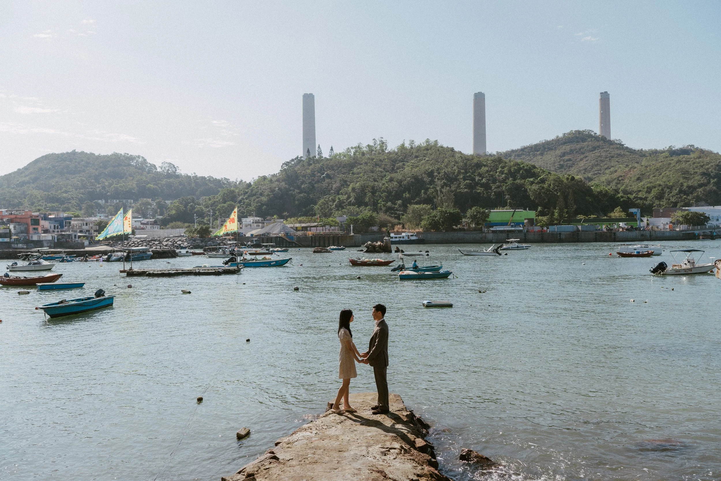 A romantic couple holding hands and standing on a stone pier by the water with boats and a hillside in the background.