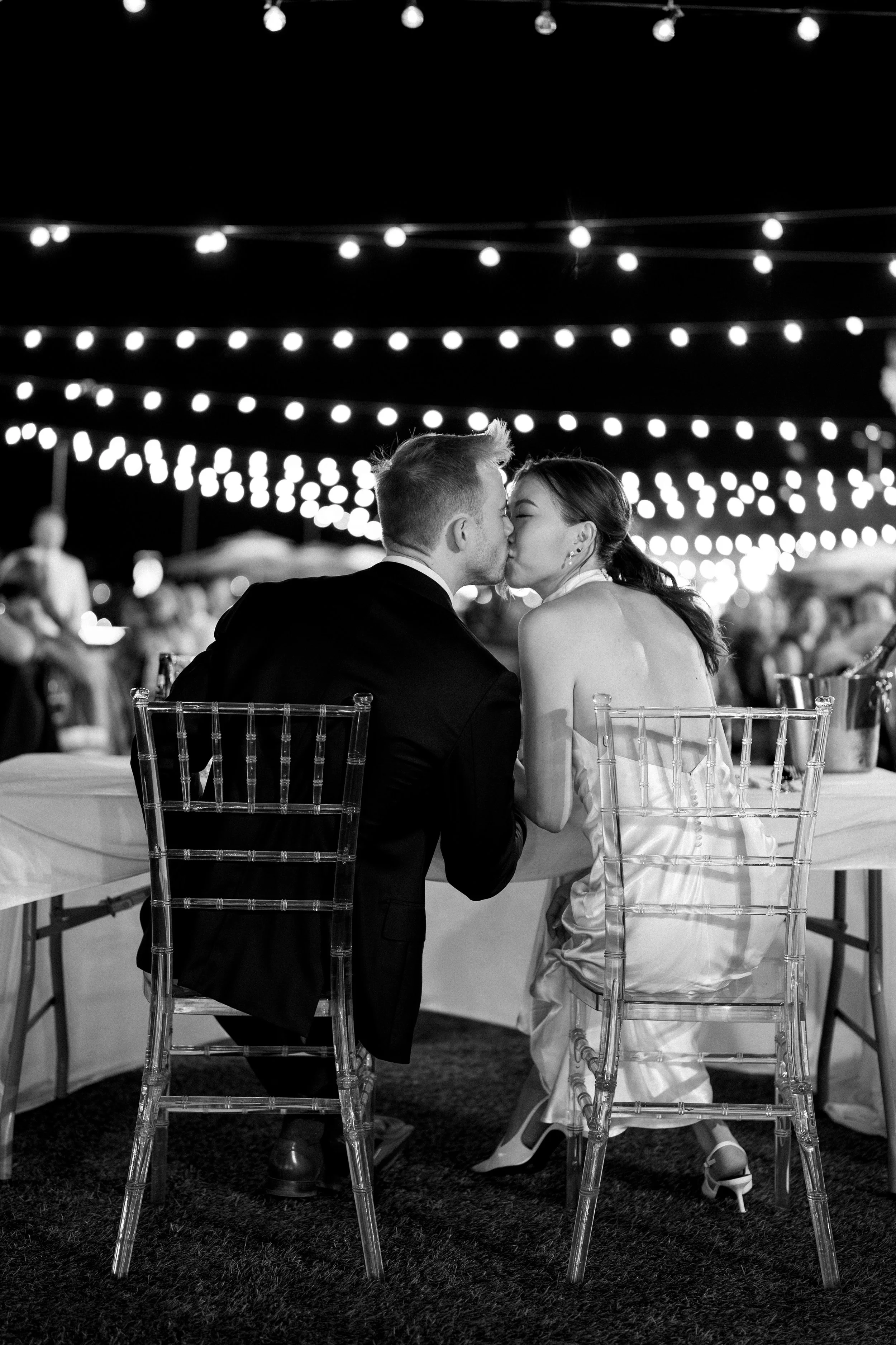 Black and white photo of a couple kissing at night during a celebration or event, with string lights overhead and a blurred crowd in the background.