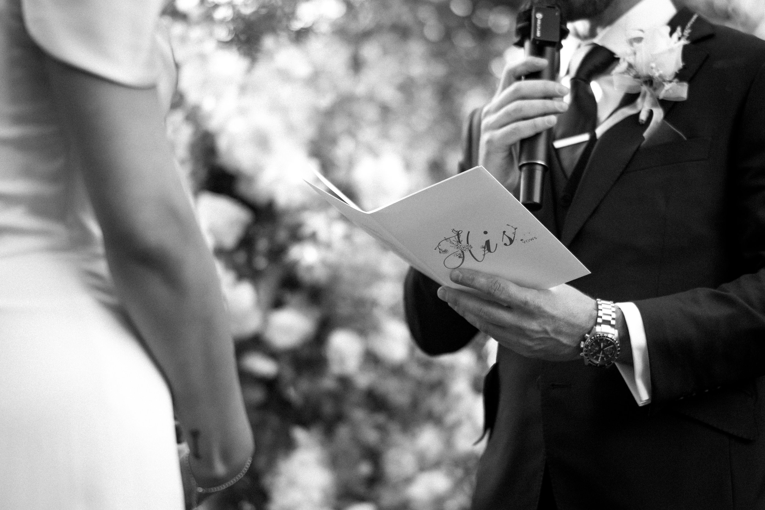 A man in a suit holding a microphone and a booklet during a wedding ceremony, with a woman in a wedding dress visible on the left side of the image.