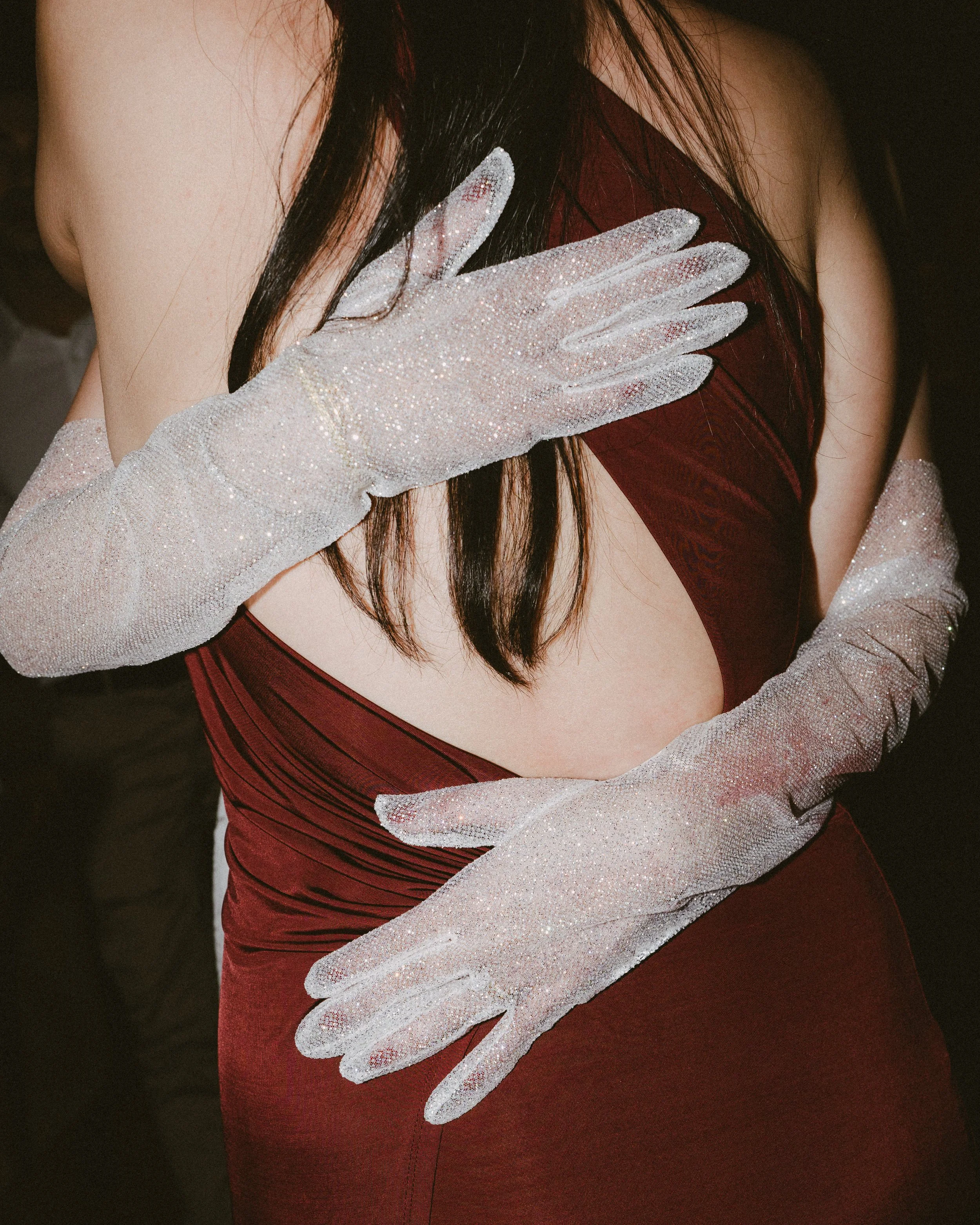 Close-up of a woman's hand with glittery white gloves resting on her chest, wearing a deep red dress.