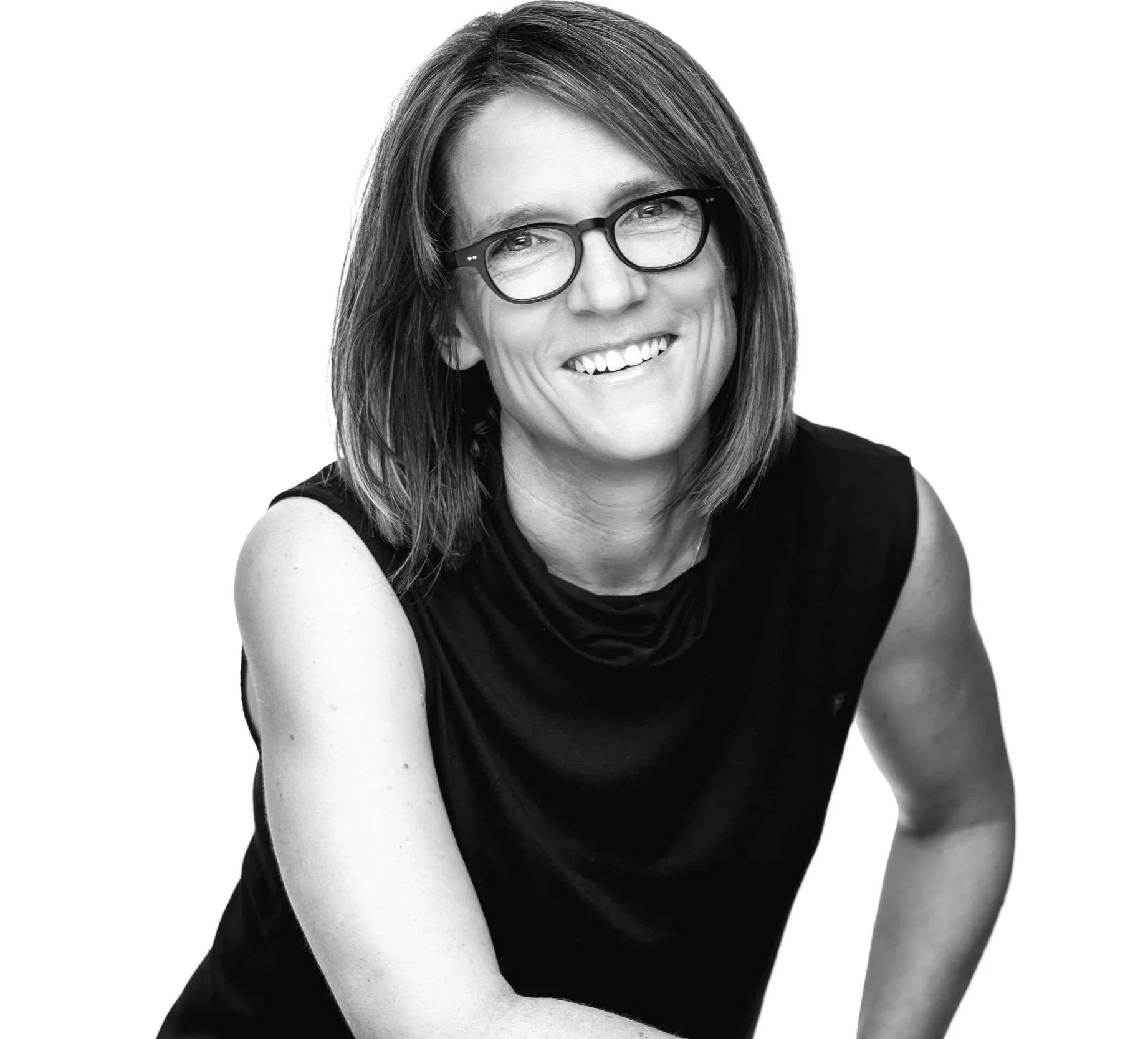 Black and white portrait of a woman wearing glasses, smiling, with shoulder-length hair, wearing a sleeveless black top, against a plain white background.