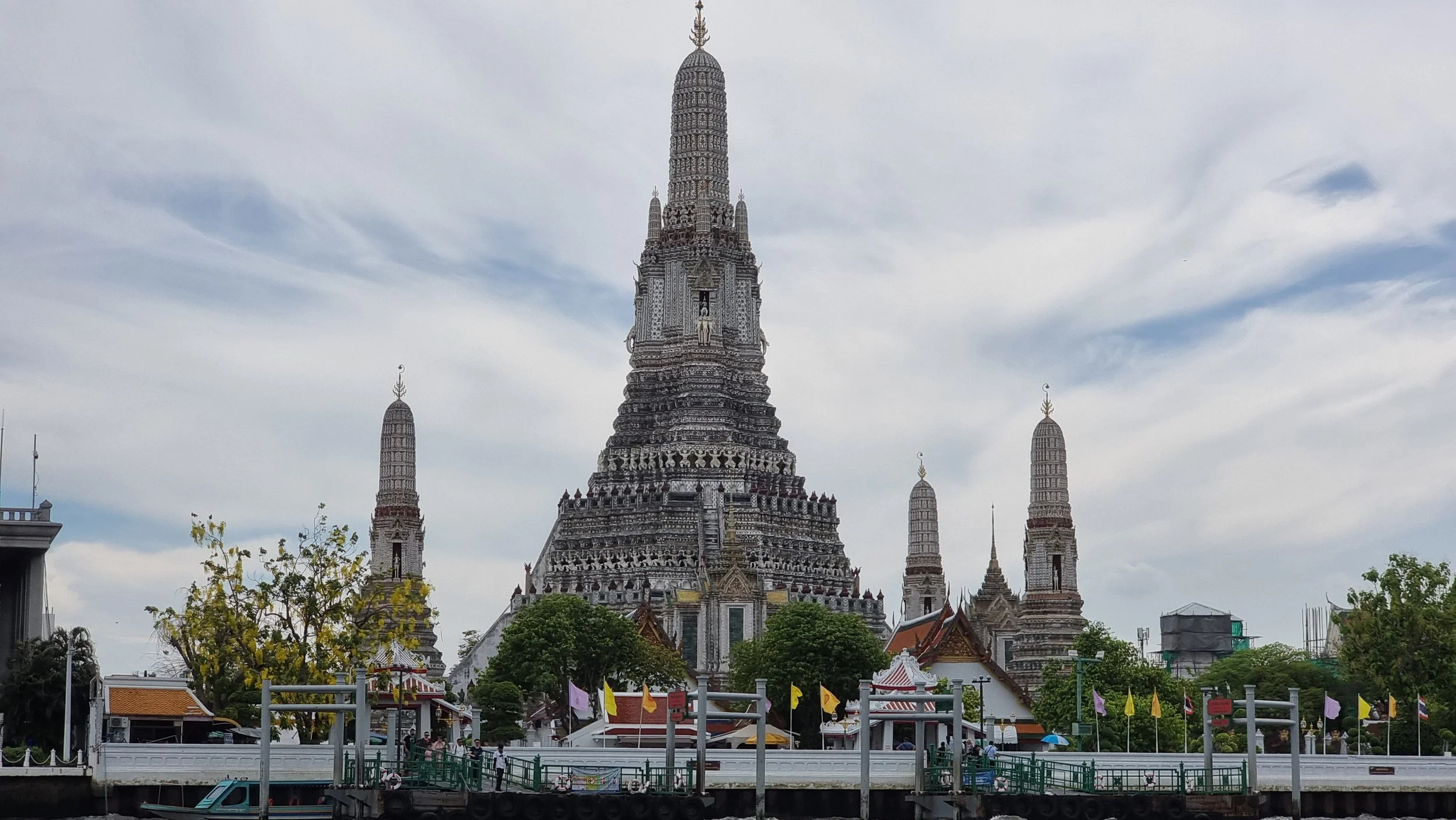 Wat Arun, a large ornate Buddhist temple with a central spire and four surrounding smaller spires, situated along the river with trees and flags in the foreground, under a partly cloudy sky.