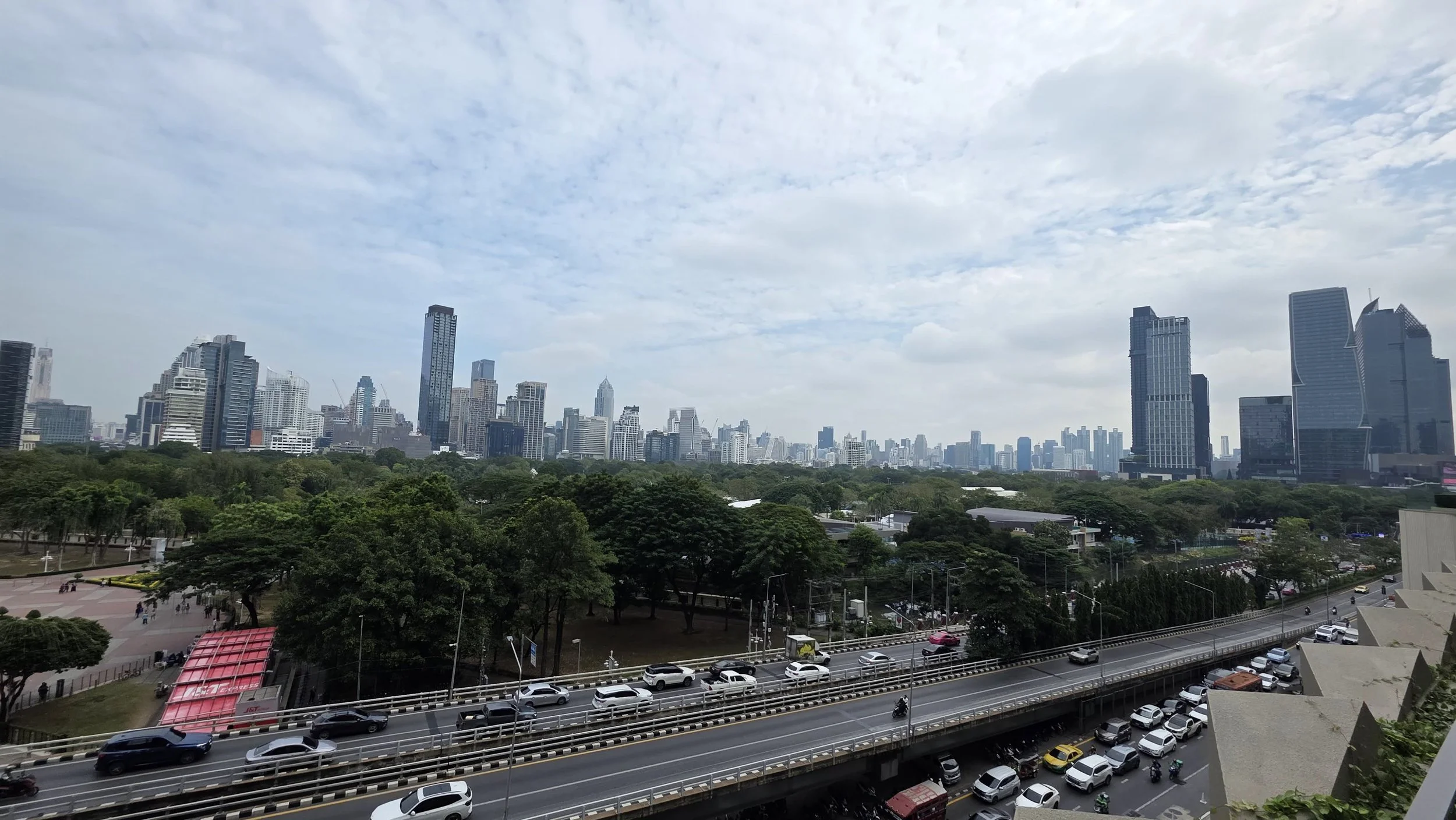 Bangkok City skyline with tall modern buildings, green trees, and a busy multi-lane road with cars and motorcycles in the foreground.