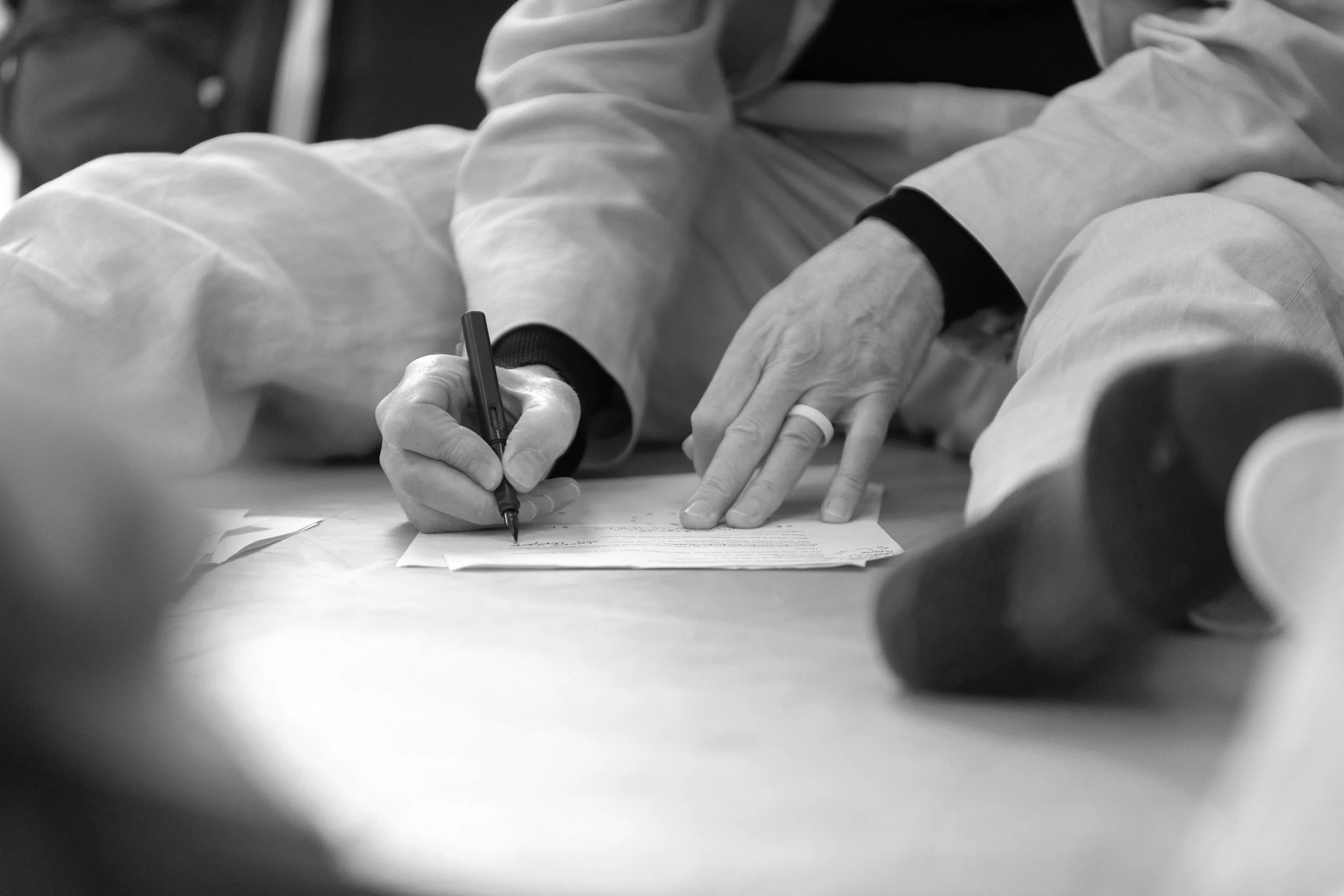A person in a white jacket signs a document on a table with a black pen, with their other hand pointing at the paper.