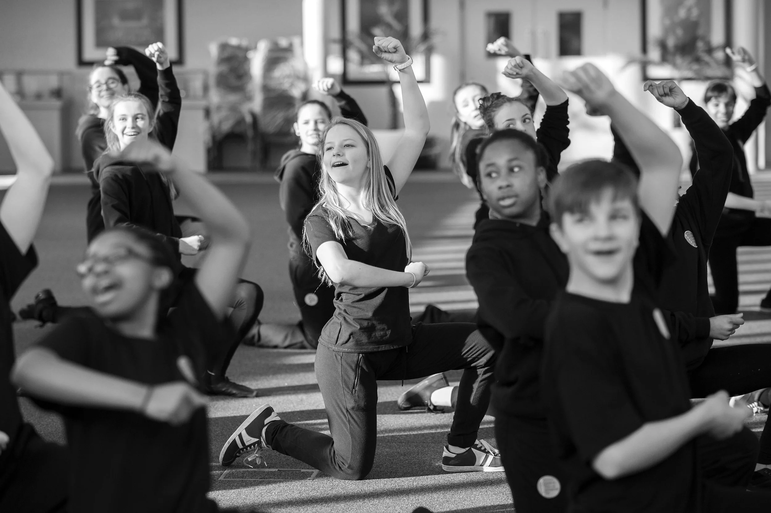 Children participating in an indoor group activity, some kneeling and raising their fists, smiling and looking in various directions, with a background of framed pictures on the wall.