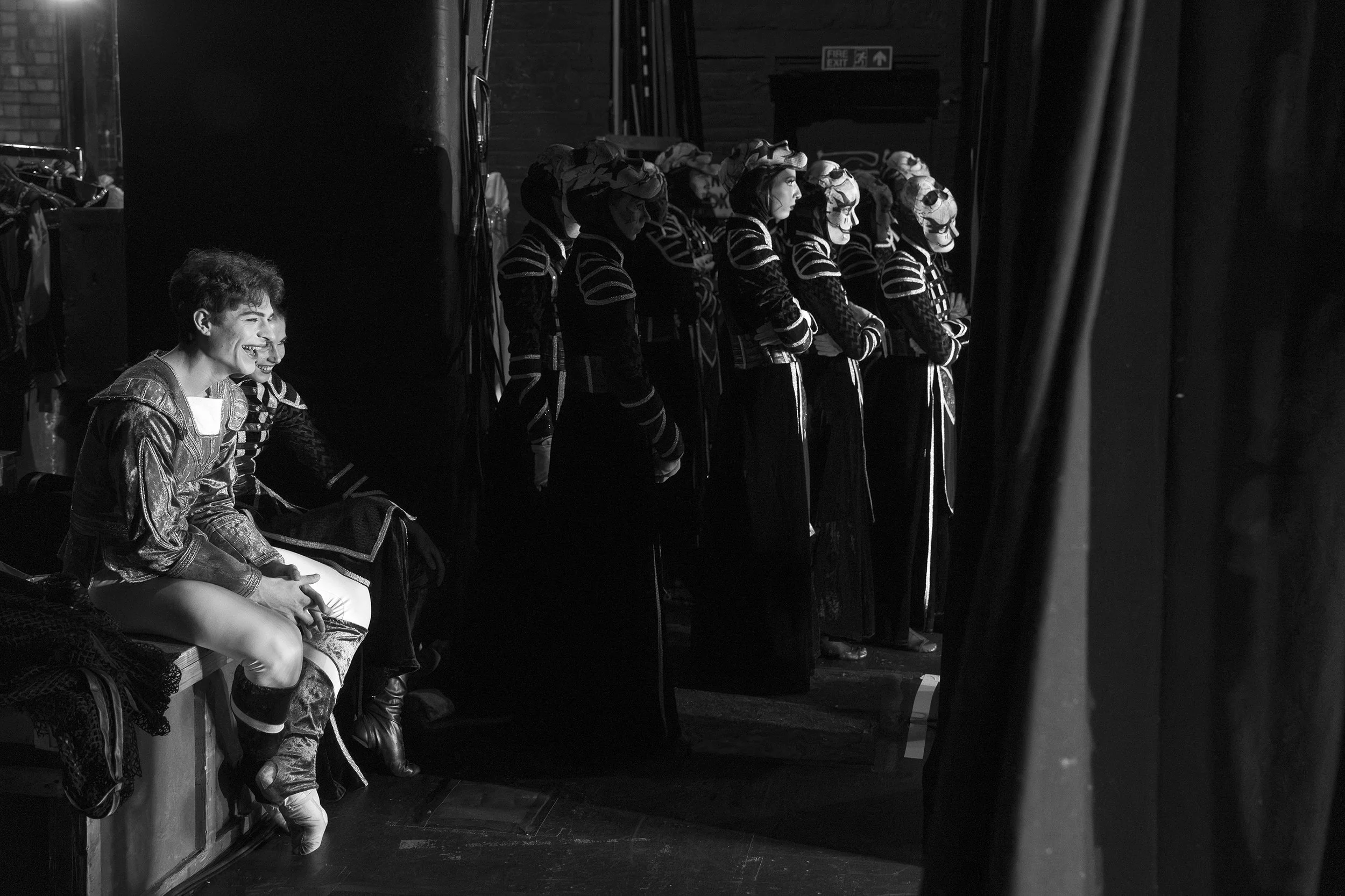 Person sitting on a bench backstage, smiling and looking towards the stage, with a line of performers dressed in costumes and masks standing nearby in the shadows, preparing for a performance.