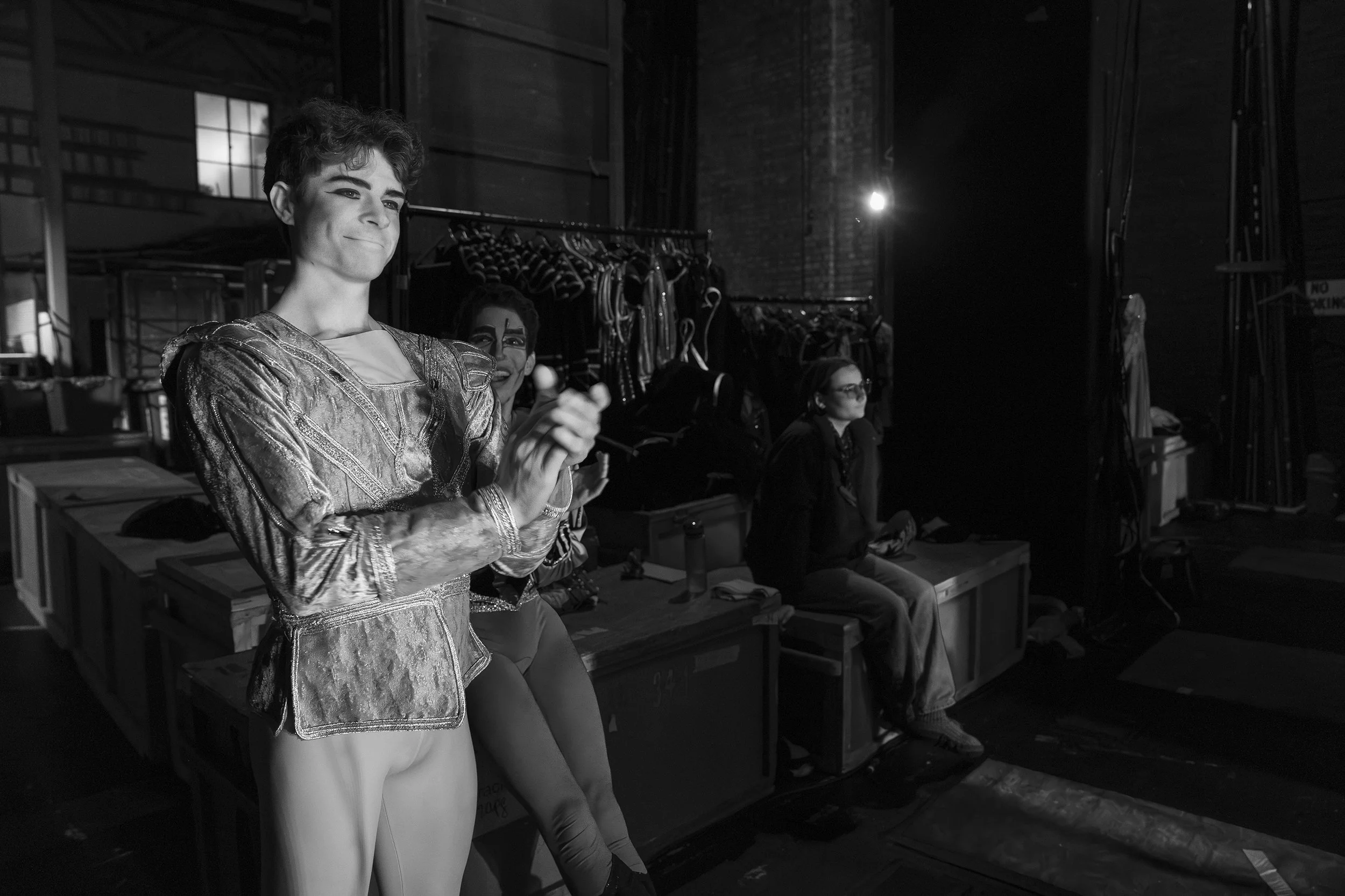 Black and white backstage photo showing two dancers in costume sitting on storage boxes, with one performer in a metallic jacket and another with face makeup, and a woman in dark glasses sitting nearby.