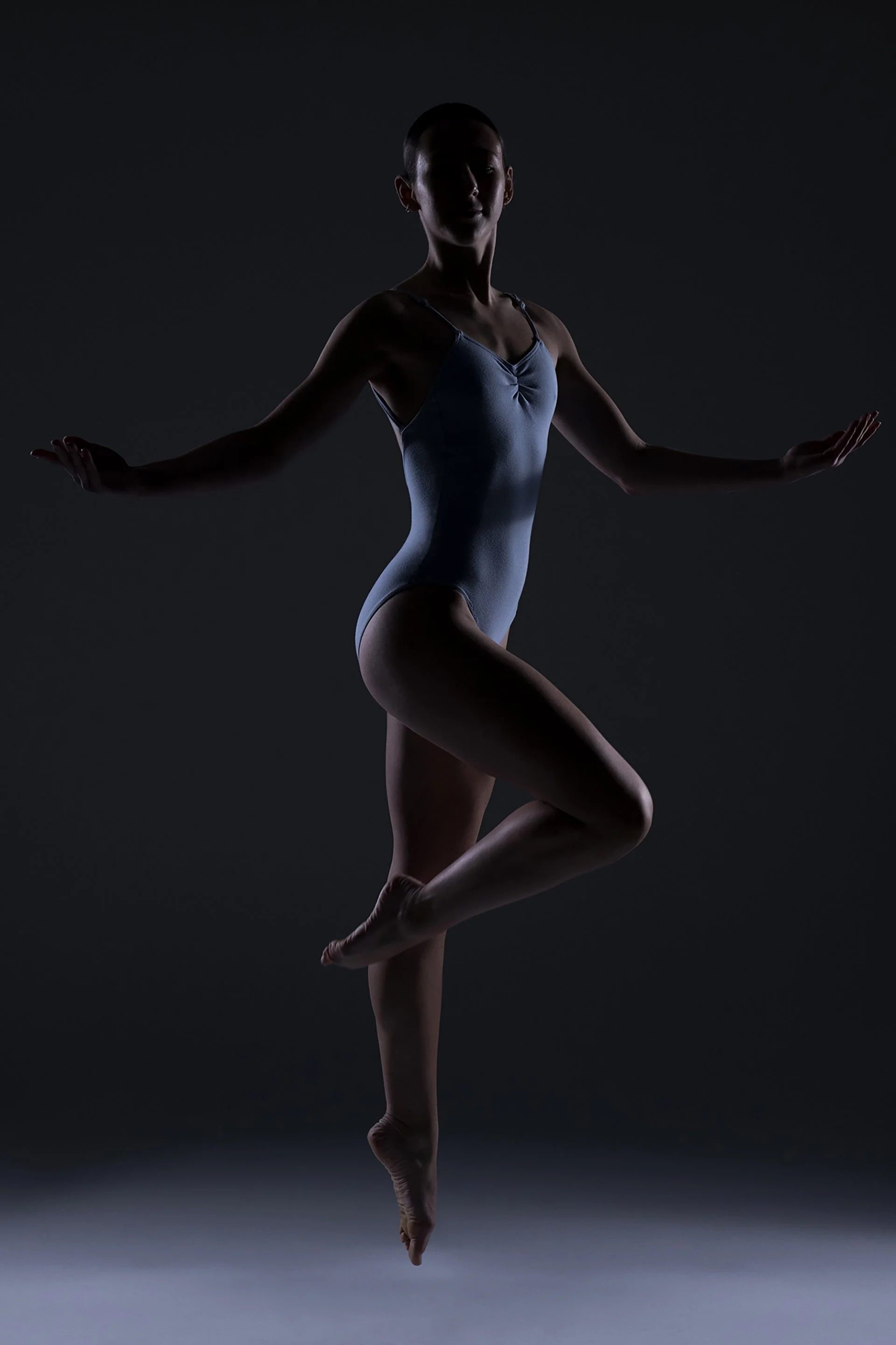Silhouette of a female gymnast performing a balance pose with one leg raised and arms extended sideways, against a dark background.