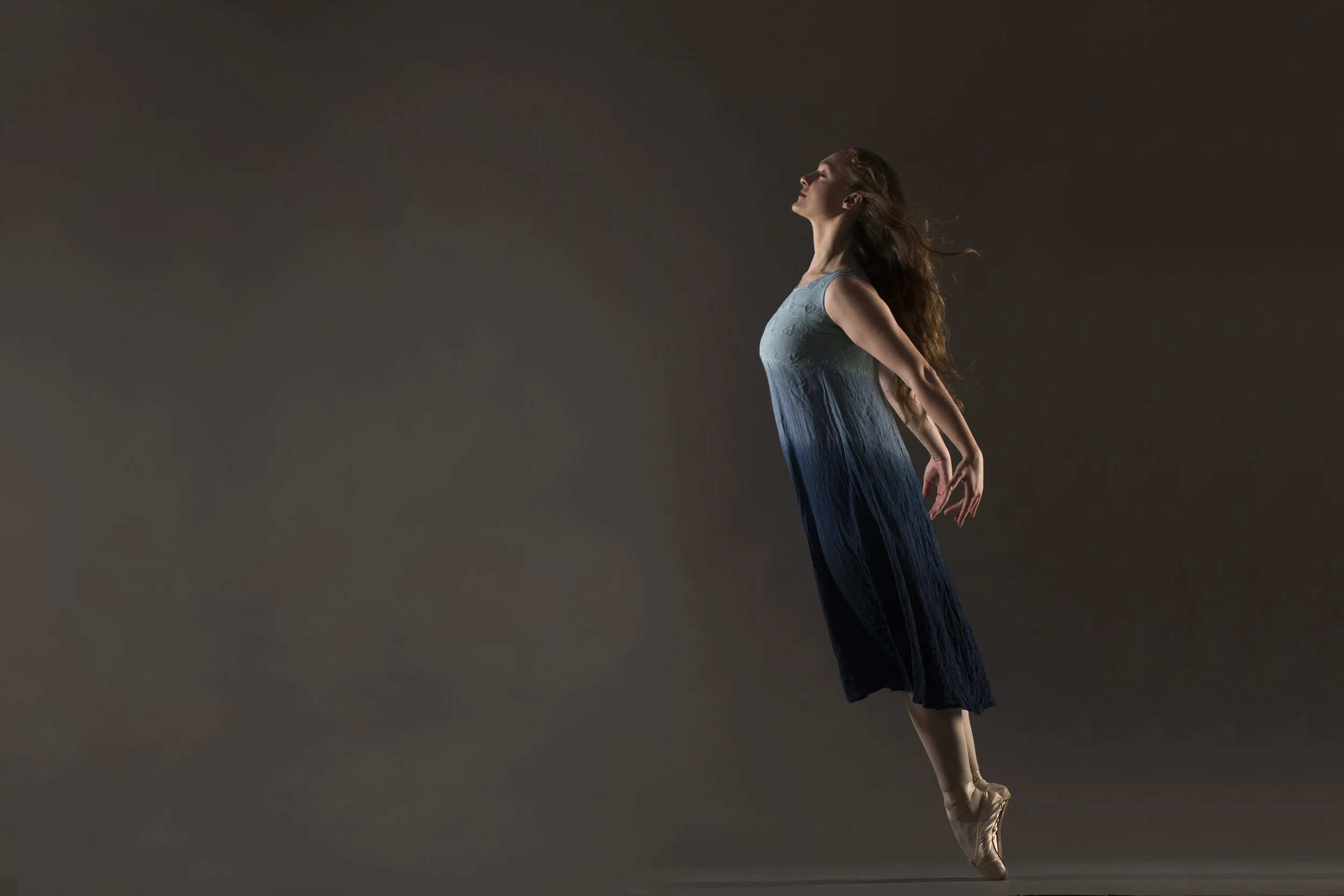 A ballerina in a blue gradient dress performing a jump with her arms and legs extended, hair flowing, against a dark background.