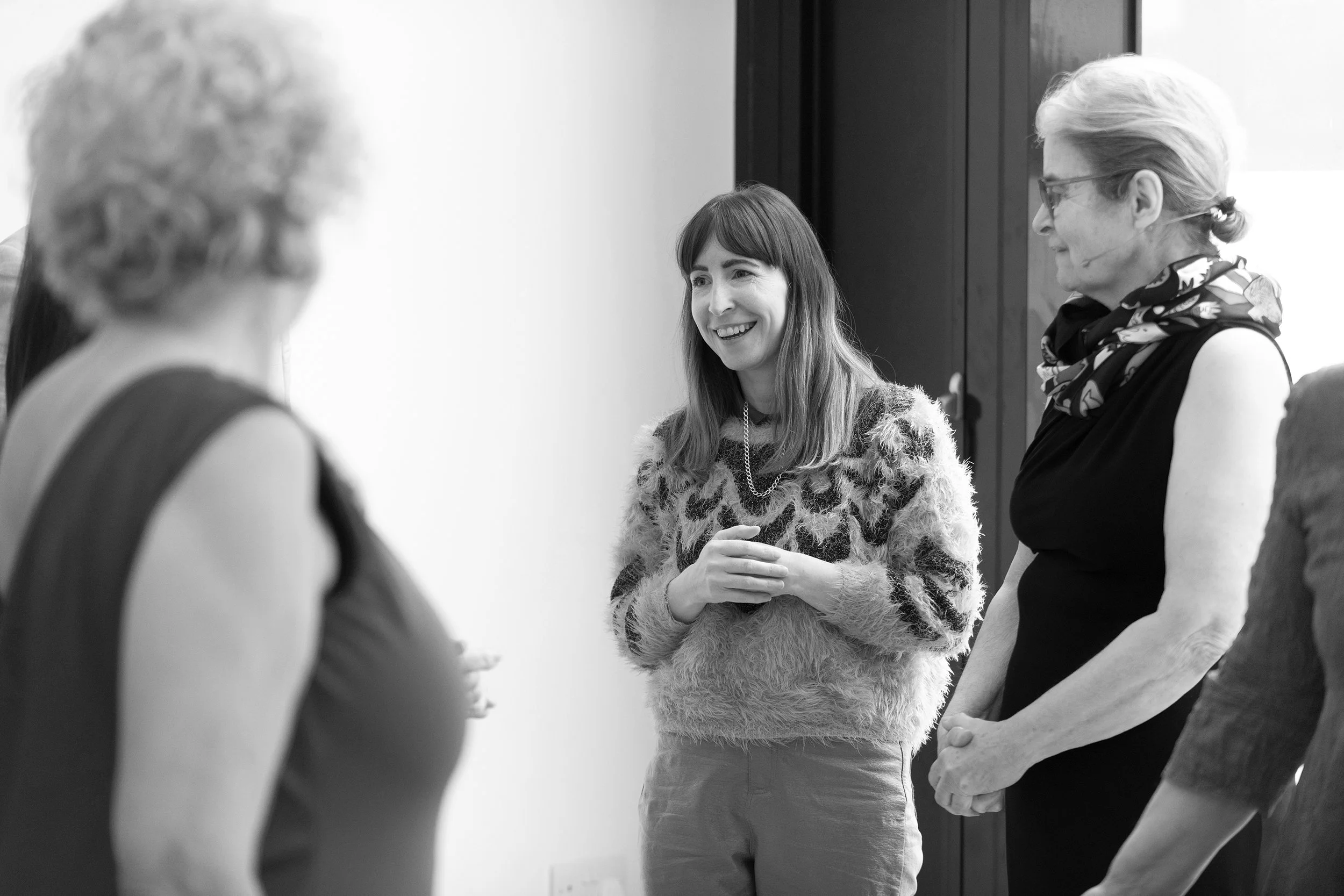 Four women engaging in conversation indoors, with one woman smiling, wearing a patterned sweater, and the others with glasses and scarves.
