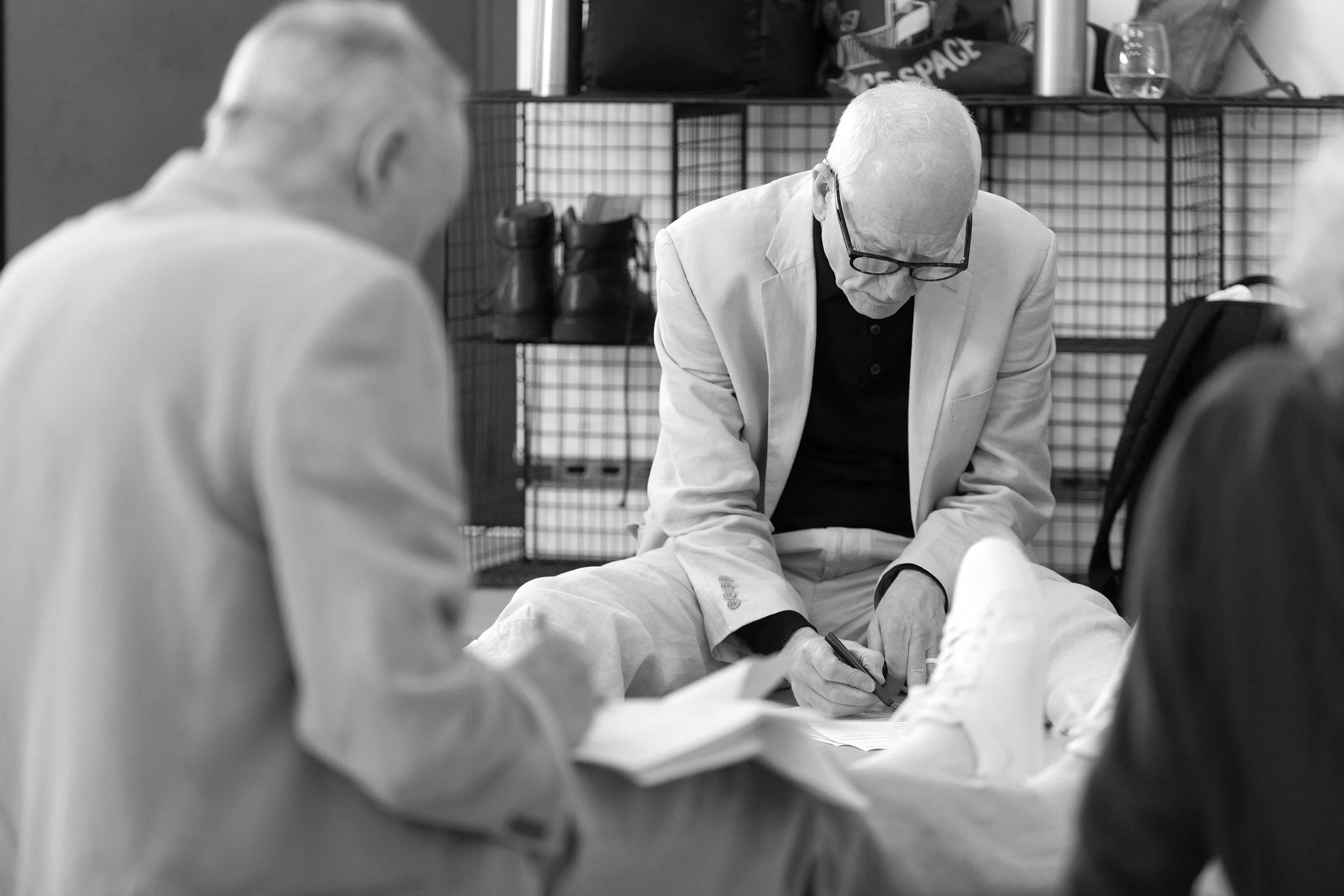 Black and white photo of an elderly man with glasses and a bald head, sitting on the floor, writing on a piece of paper. Other people are partially visible around him, and there are shelves with shoes and bags in the background.