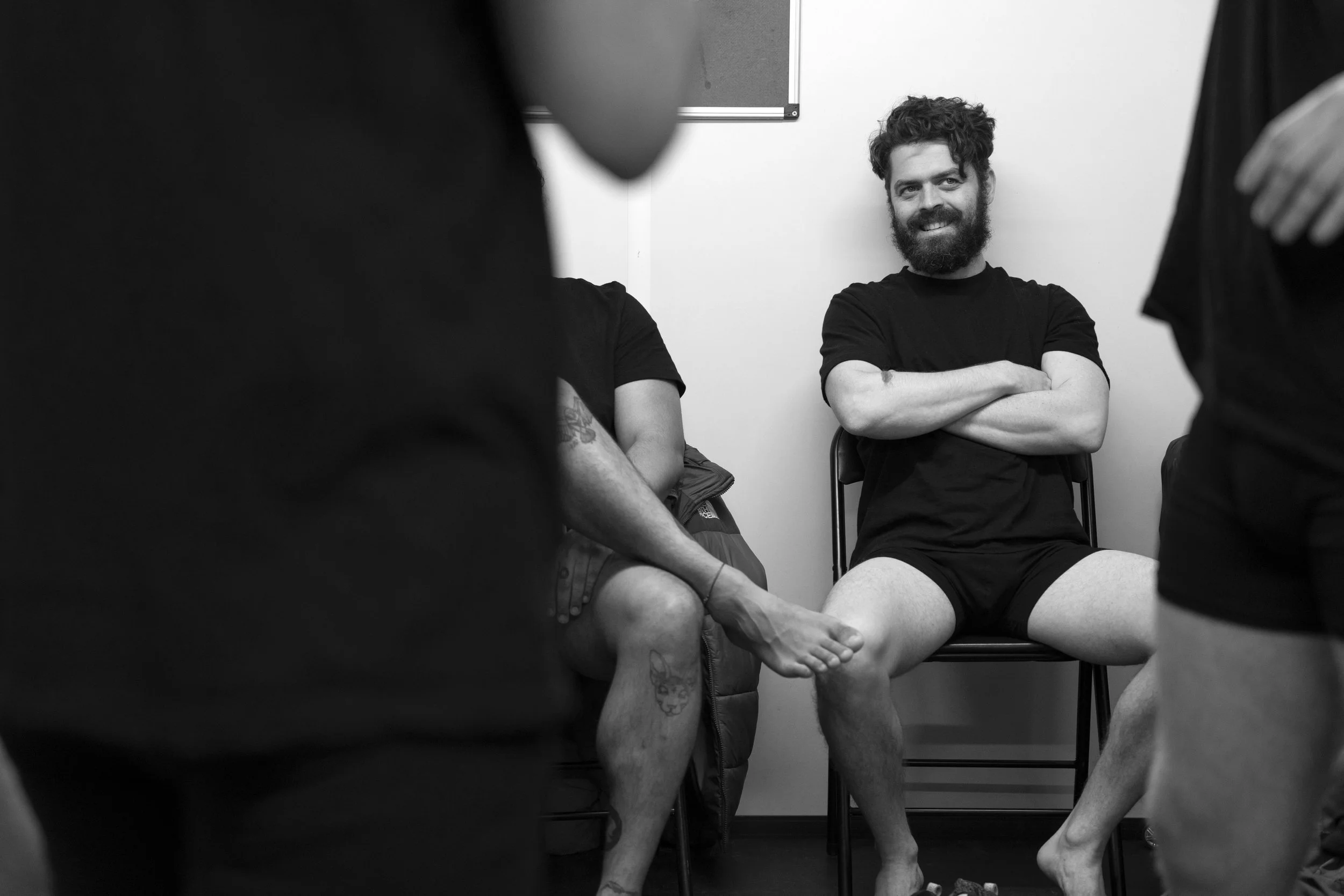 A group of people sitting in a dressing room getting ready to go on stage, with one man smiling, wearing a T-shirt and shorts, crossing his arms and legs, and others partially visible.