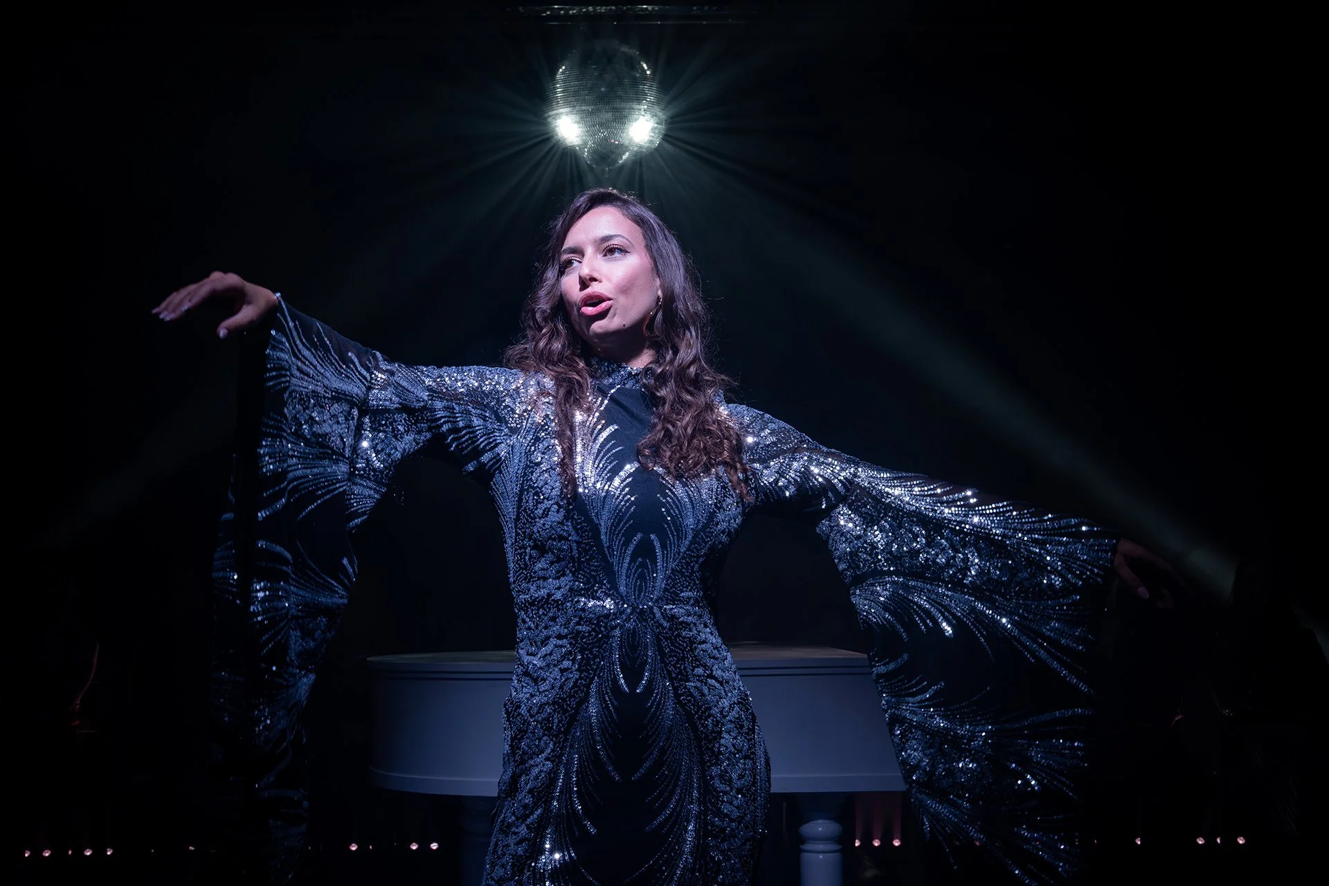 A female actor in a shiny, patterned, long dress with wide sleeves, standing under a disco ball with light reflections, posing with arms outstretched on a dark stage.