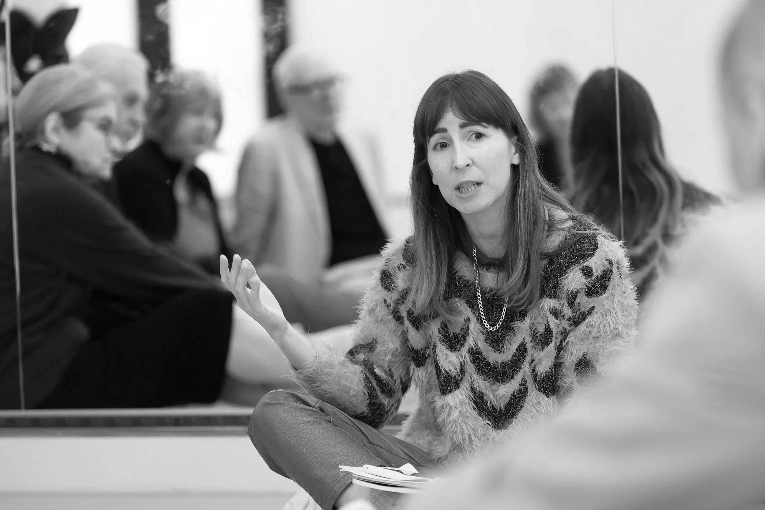 A woman with shoulder-length hair wearing a patterned sweater and a chain necklace, speaking and gesturing with her hand, sitting on the floor in front of a mirror, with several blurred individuals seated behind her.