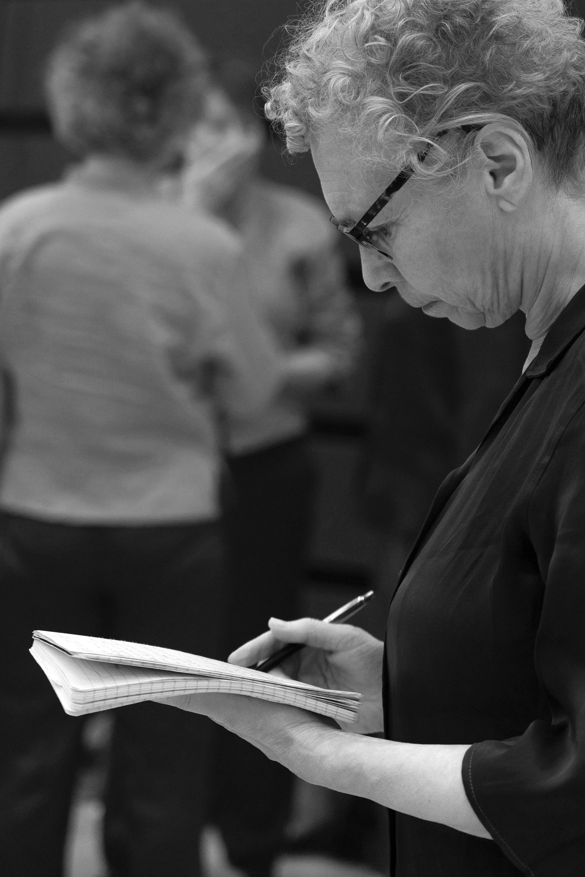 A woman with curly hair and glasses is taking notes in a notepad with a pen while standing in a busy indoor setting, with people in the background.