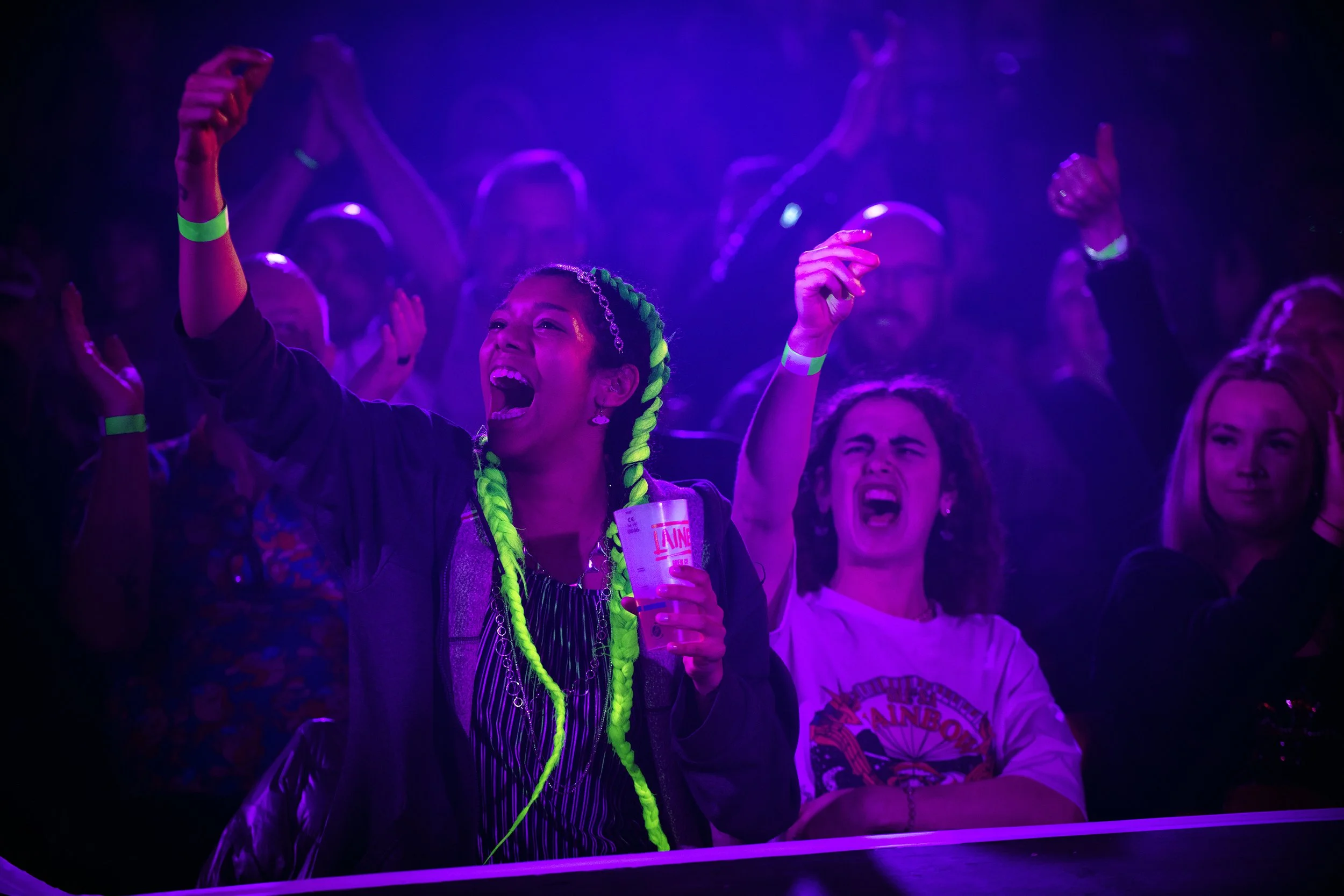 People enjoying a Fringe festival performance are  illuminated by purple and blue lights, some wearing neon accessories, and cheering with their hands in the air.