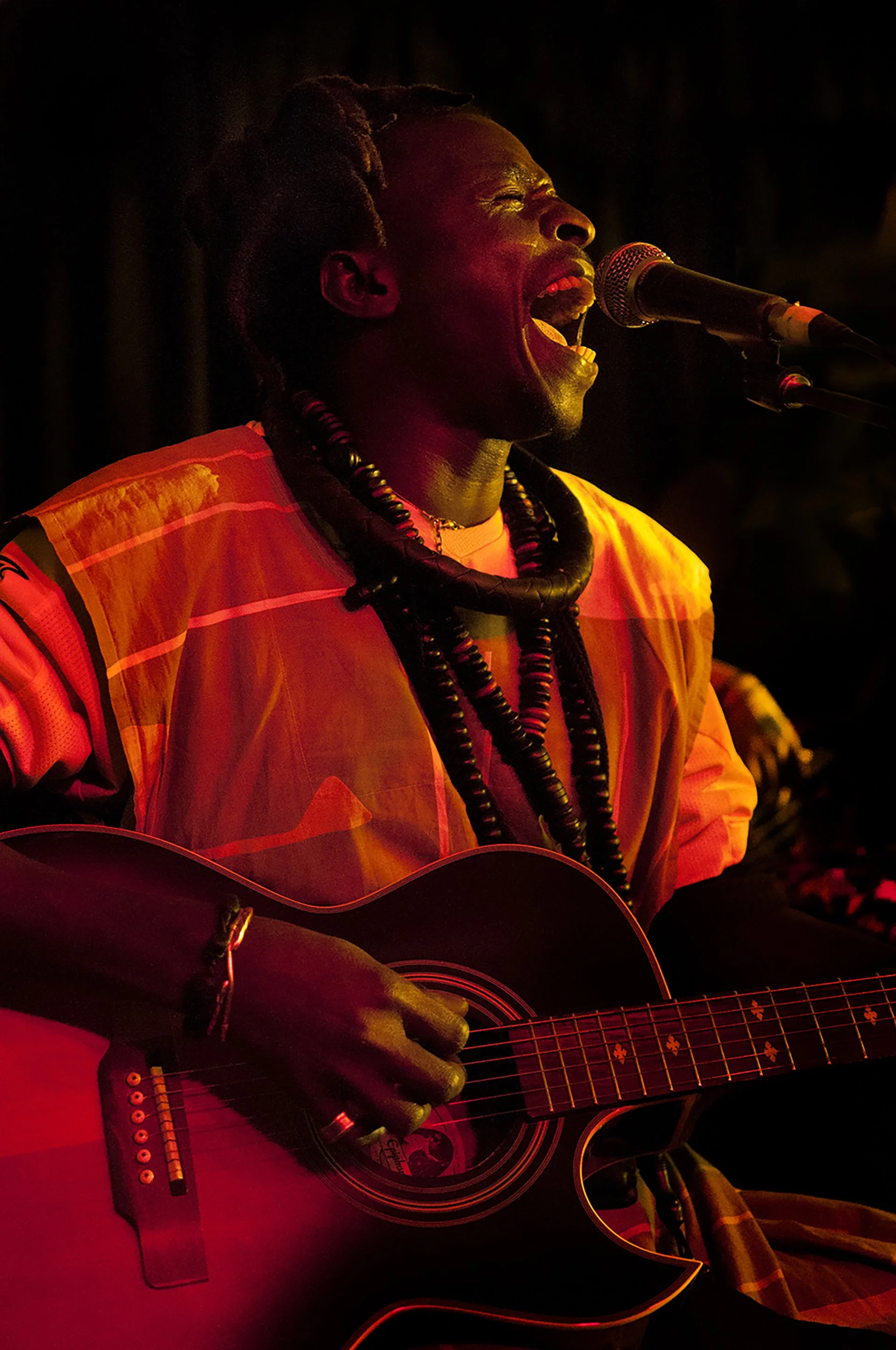 A person with dark skin and dreadlocks singing passionately into a microphone while playing an acoustic guitar. They are wearing traditional jewelry and an orange, yellow, and red garment.