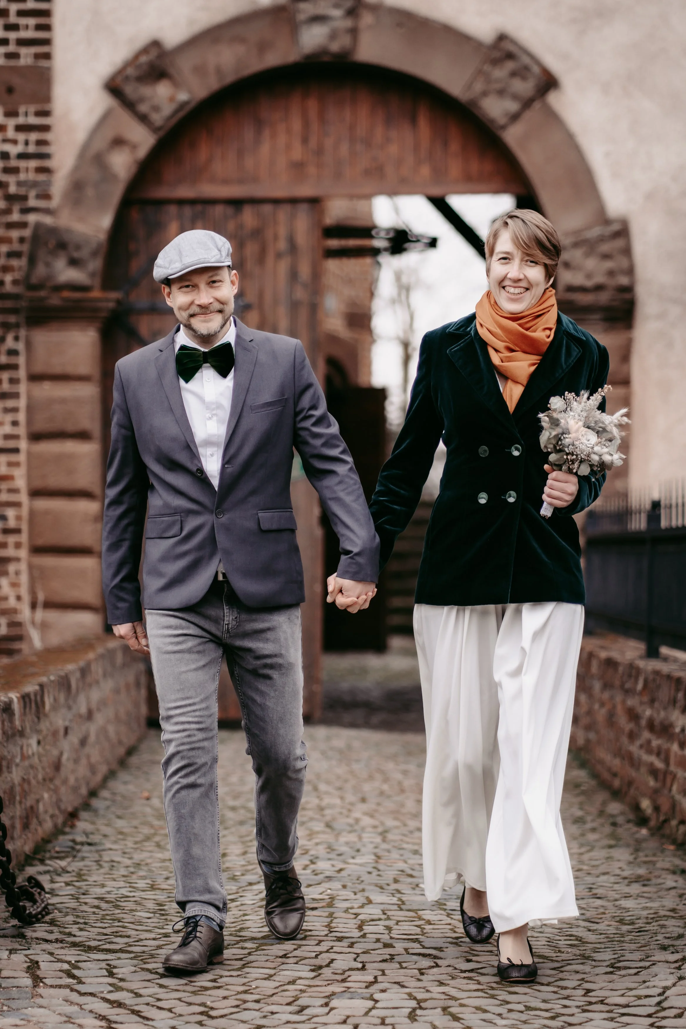 Ein lächelndes Paar in eleganter Kleidung hält Händchen und spaziert auf einem gepflasterten Weg vor einem alten Gebäude.