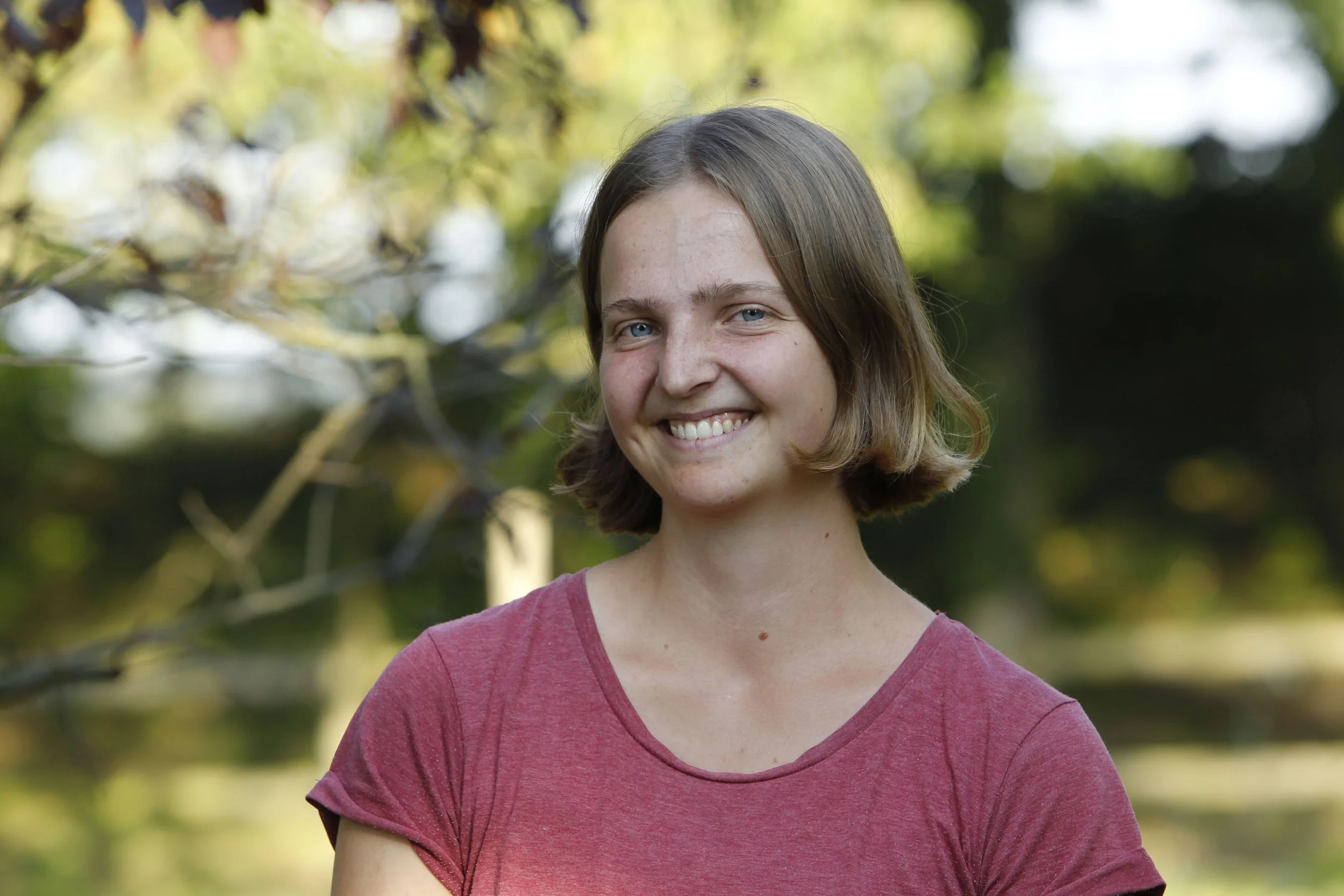 Eine Frau mit kurzem, braunem Haar lächelt in die Kamera, trägt ein rotes T-Shirt und steht im Freien vor einem verschwommenen Hintergrund aus Bäumen und Natur.