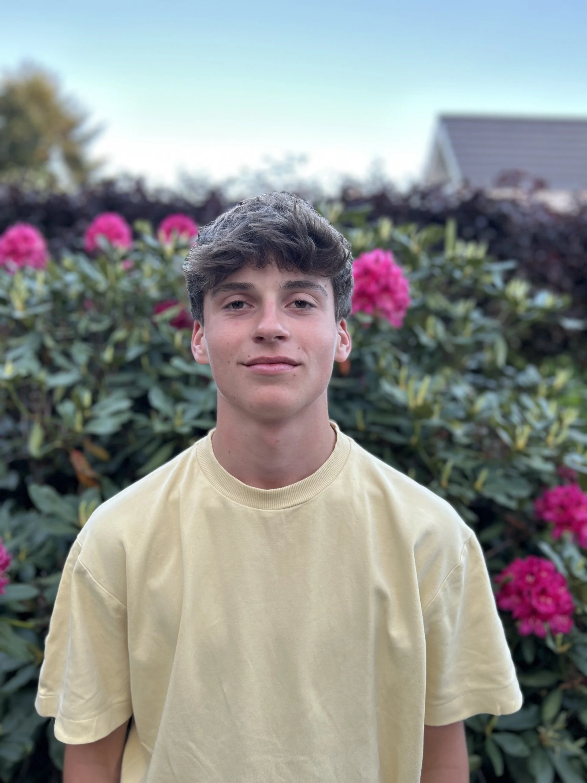 Jongeman met lichtgele T-shirt posed voor een bloeiende struik met roze bloemen in een tuin onder een blauwe hemel.