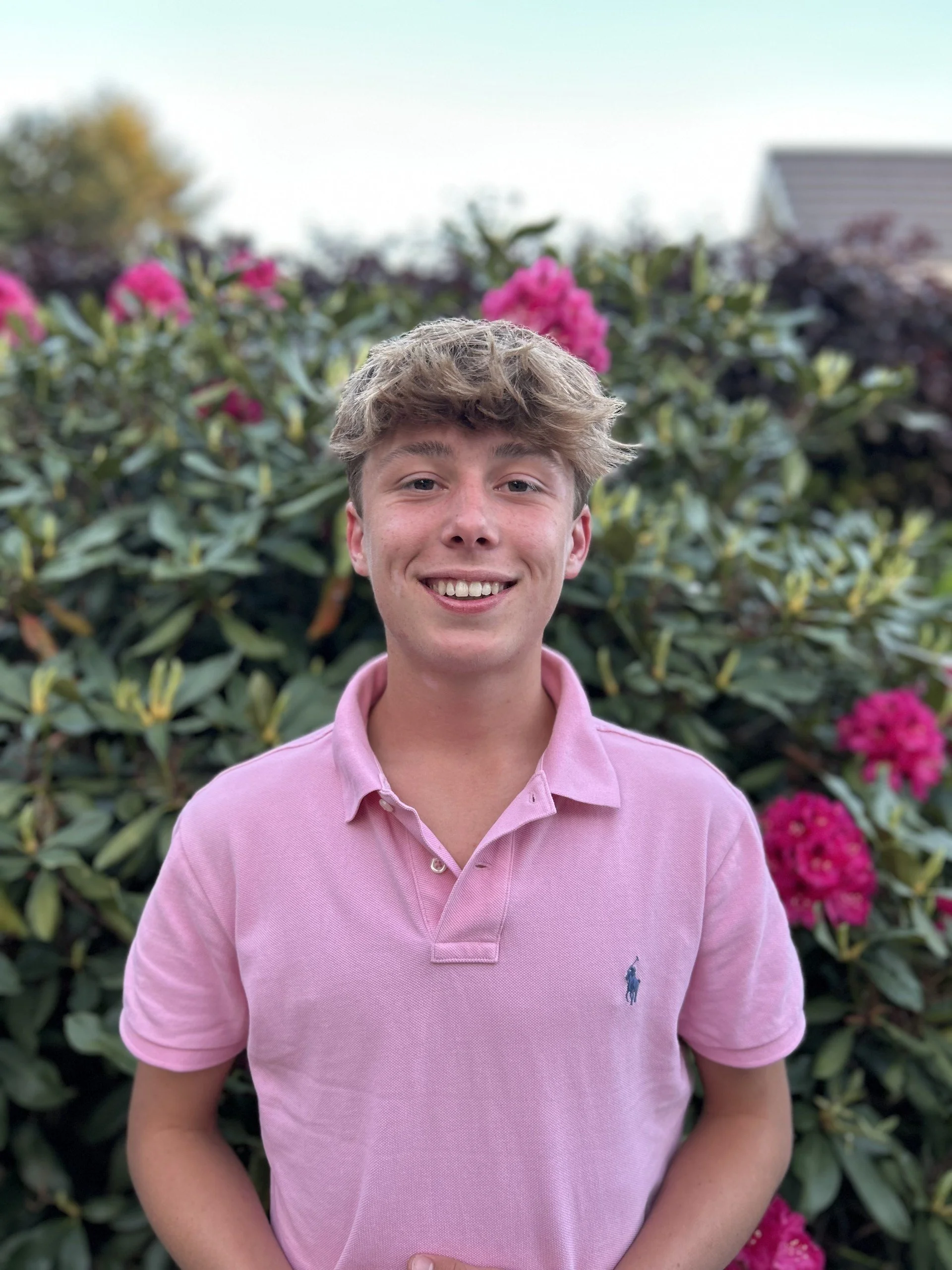 Jongen met lichtbruine haar en een glimlach, dragend een roze polo, voor een heg met roze bloemen.