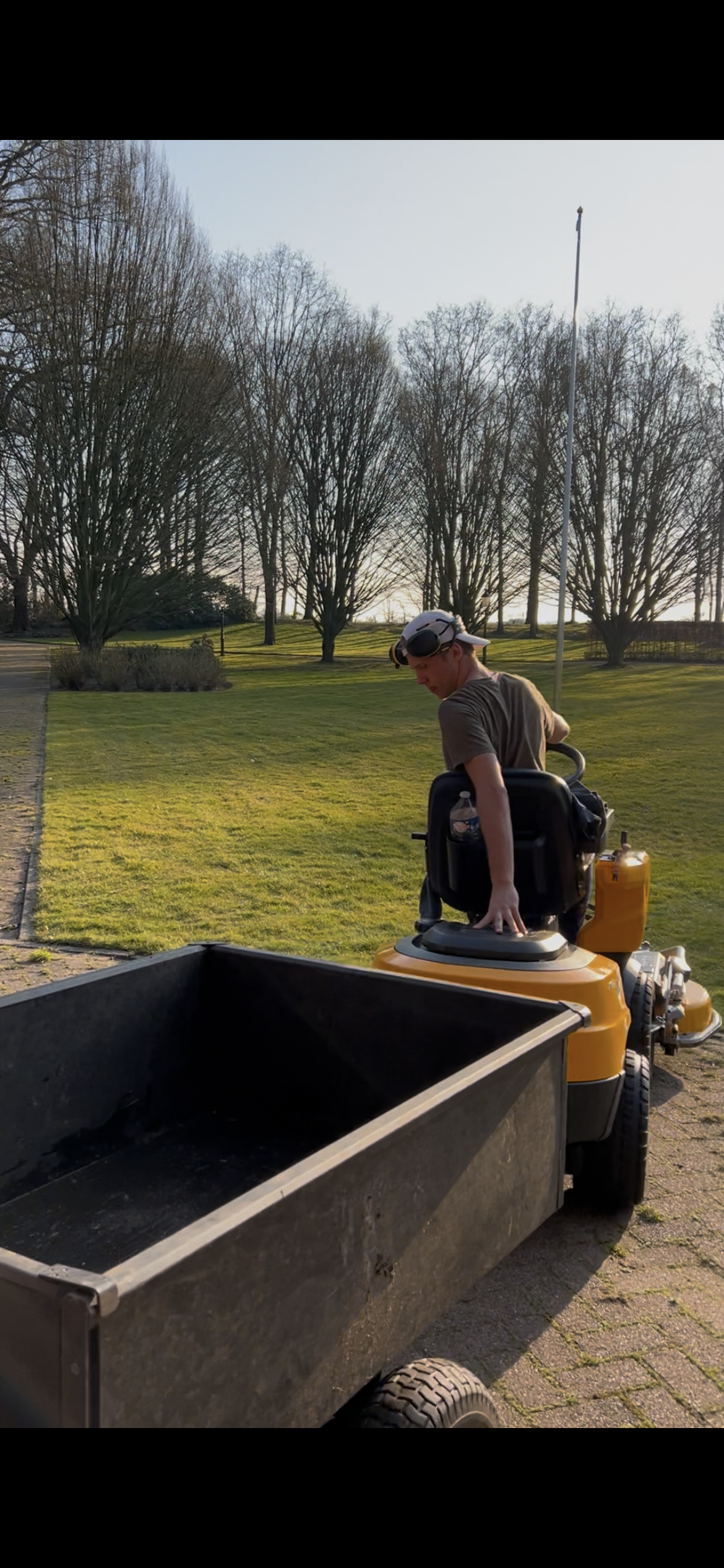 Man op een zitmaaier op het gras met een aanhangwagen, omgeven door bomen.