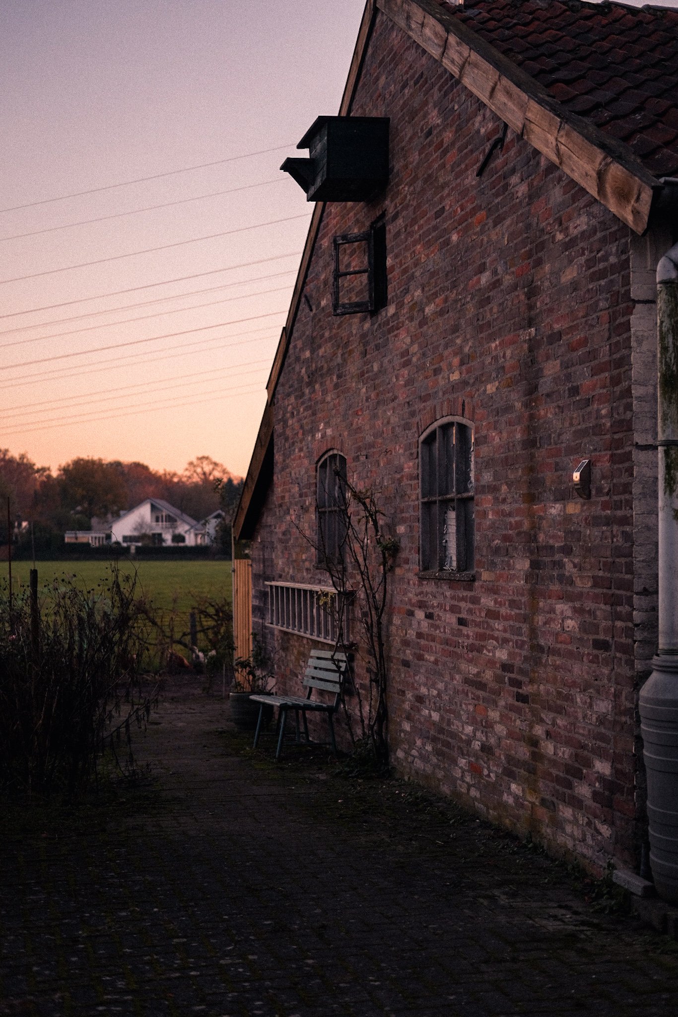 Old brick building with arched windows, outdoor bench, and green landscape at sunset.