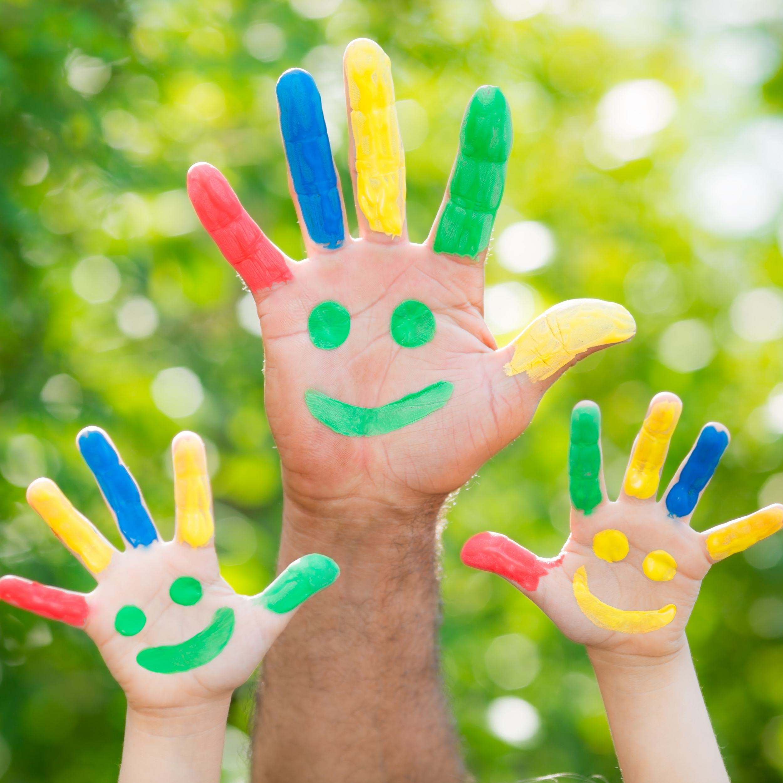 Mano con dita dipinte in diversi colori, pollice, indice, medio, anulare e mignolo, con faccia sorridente disegnata con vernice verde sulla pelle. Sullo sfondo sfocato di foglie verdi e luce naturale.