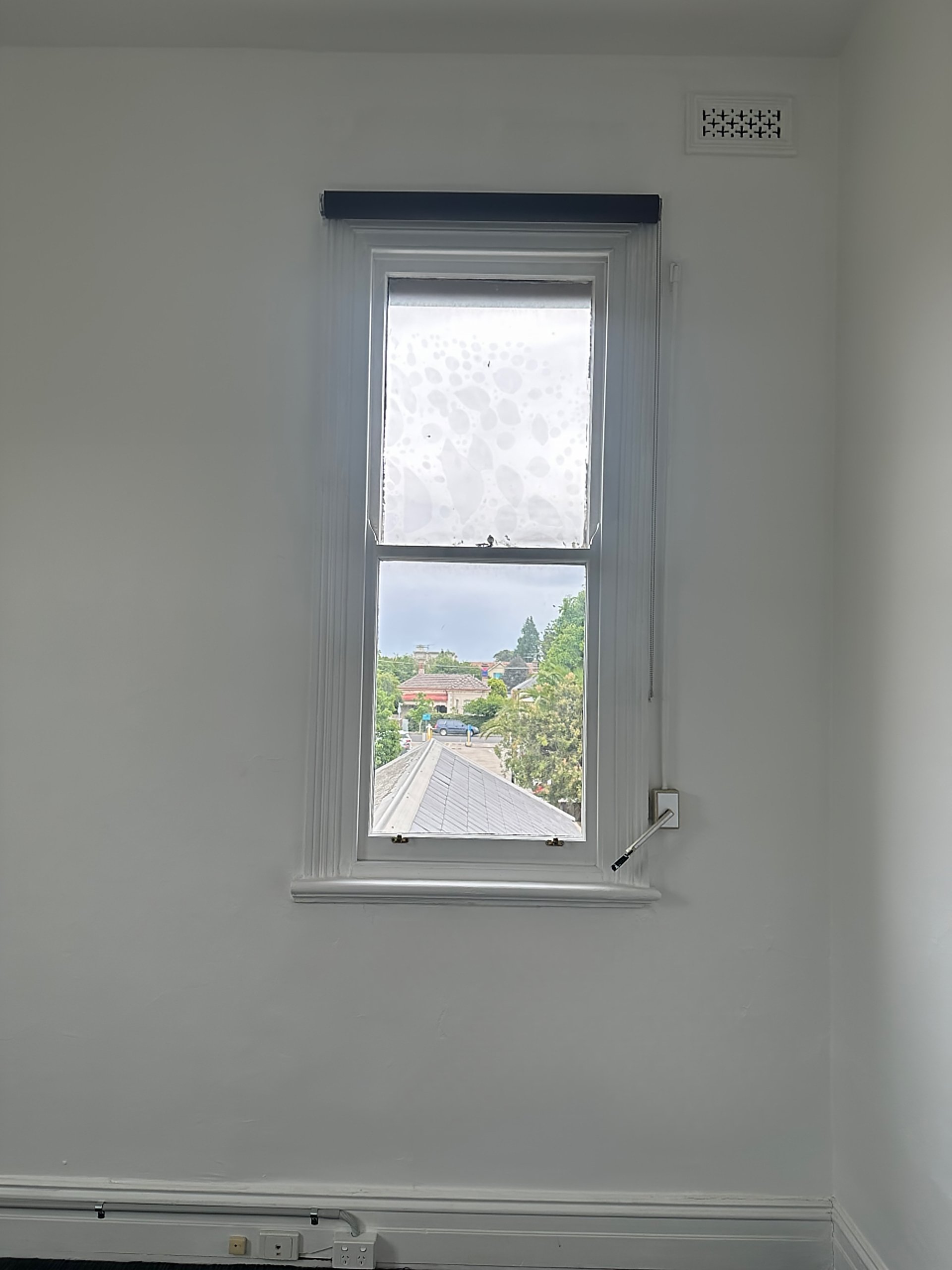 A white wall with a window divided into two parts, with one part frosted and the other clear showing a neighborhood with trees and rooftops. A black roller blind is partially rolled up at the top of the window, and a white cord hangs from the right s