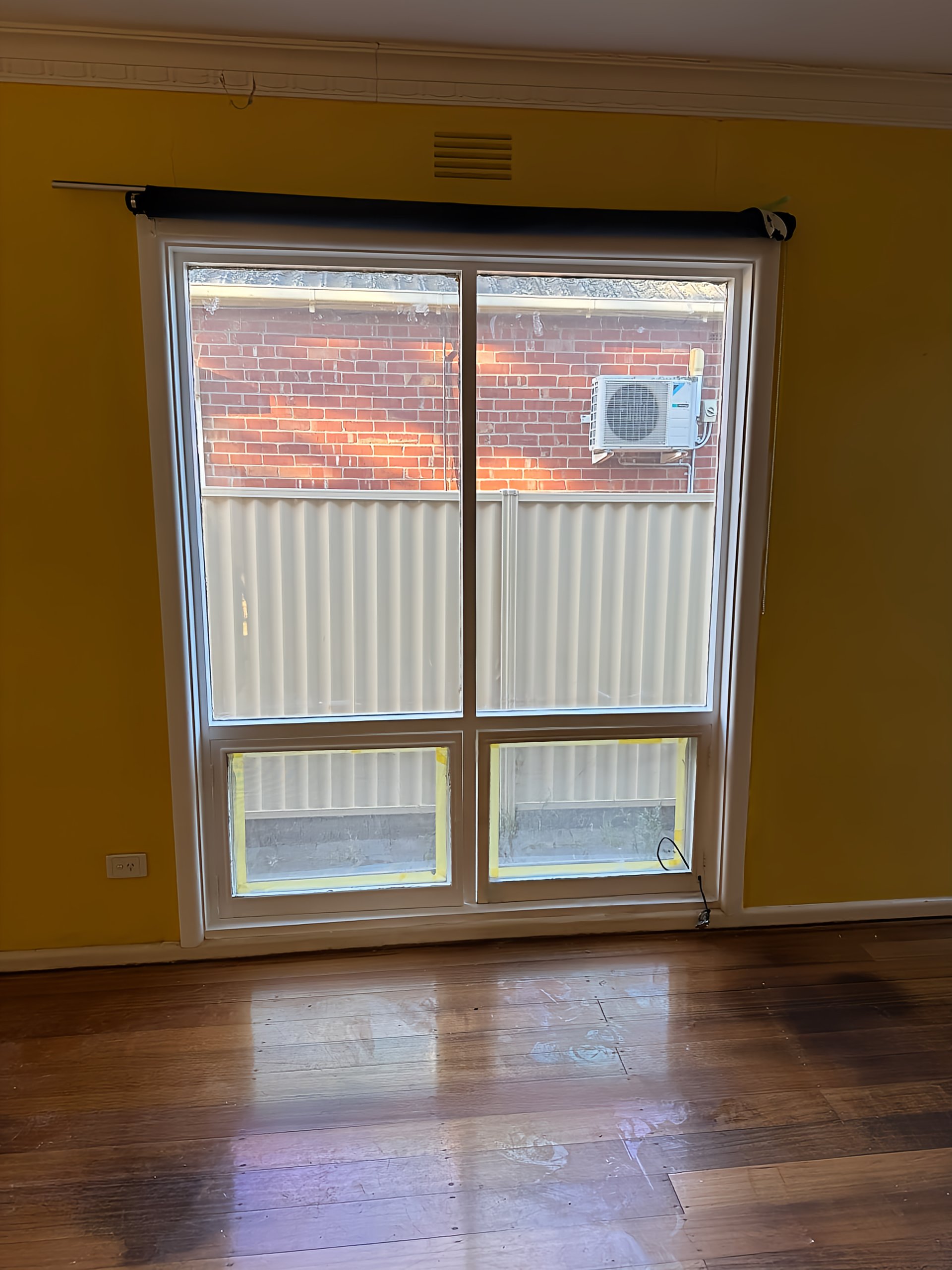Interior view of a room with wooden floor and yellow walls, showing a large window with four panes, partially covered by yellow tape at the bottom. Outside the window, there is a beige fence, a brick building, and an air conditioning unit mounted on 