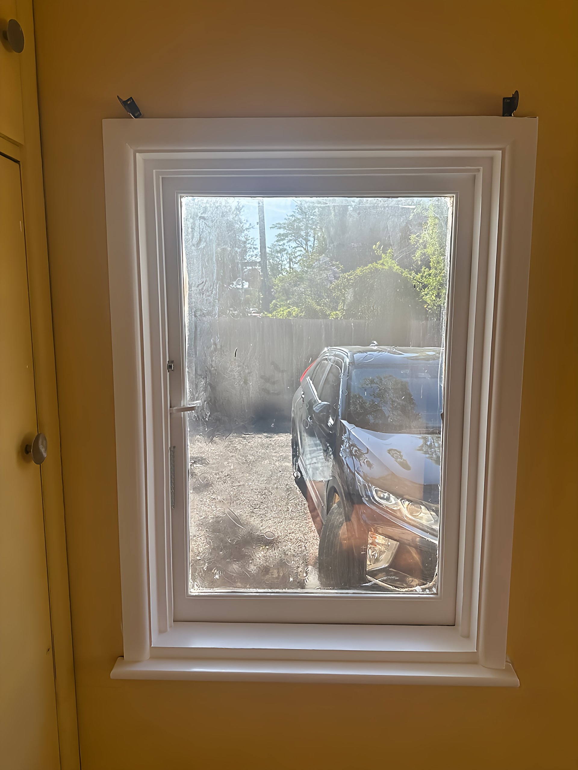 View of a black car through a dirty window with smudges and dust, outside a backyard with trees and a fence.