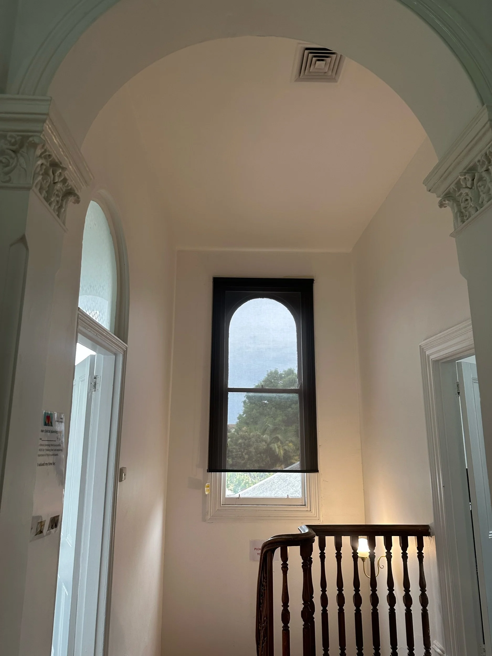 Interior of a stairway landing with a tall arched window, ornate white molding, and a wooden handrail on the staircase.