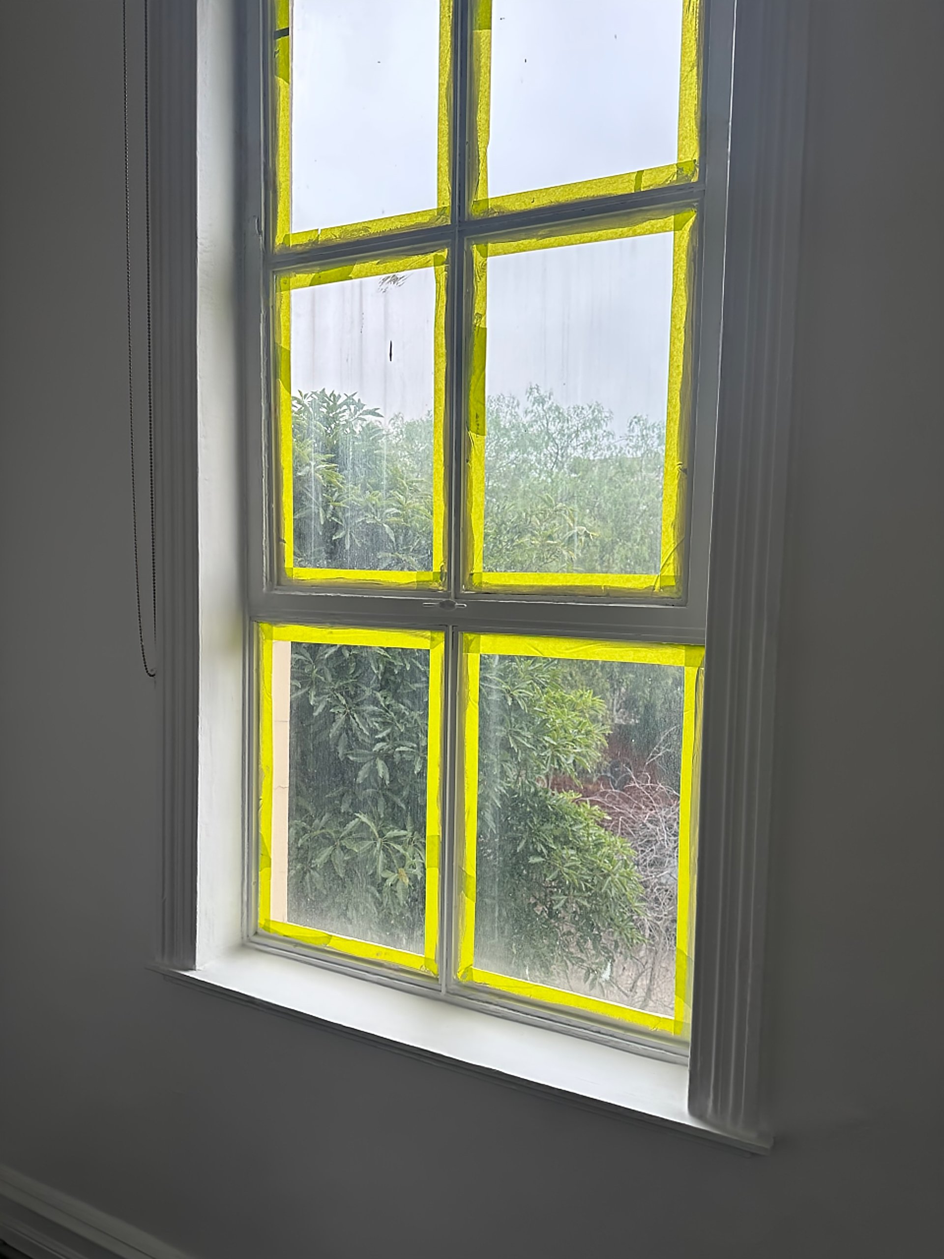 Close-up of a window with yellow painter's tape on the inside, dividing the glass into sections, with trees visible outside.