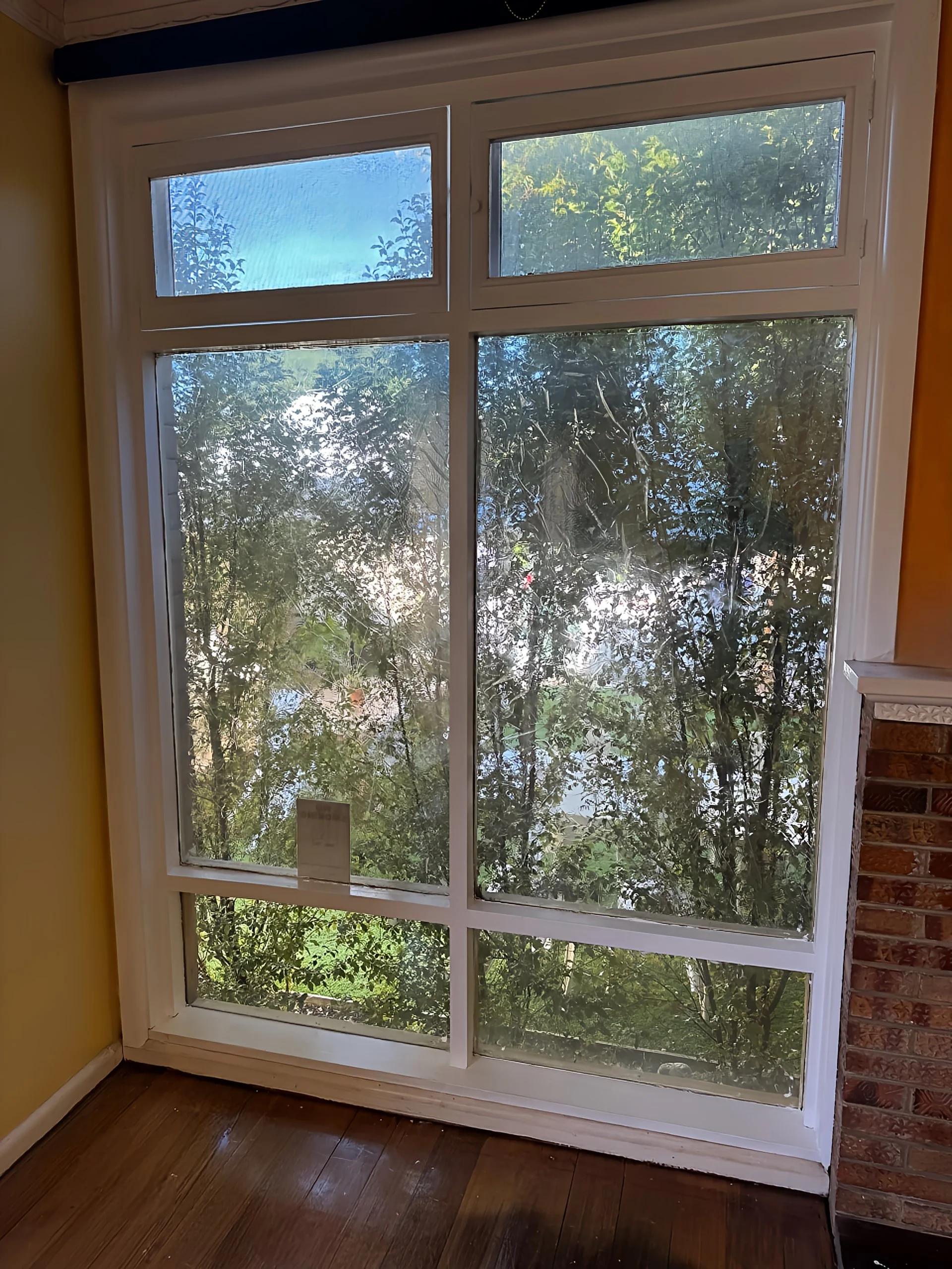 Large window with multiple panes overlooking trees and foliage outside, with a brick wall on the right and wooden floor inside.