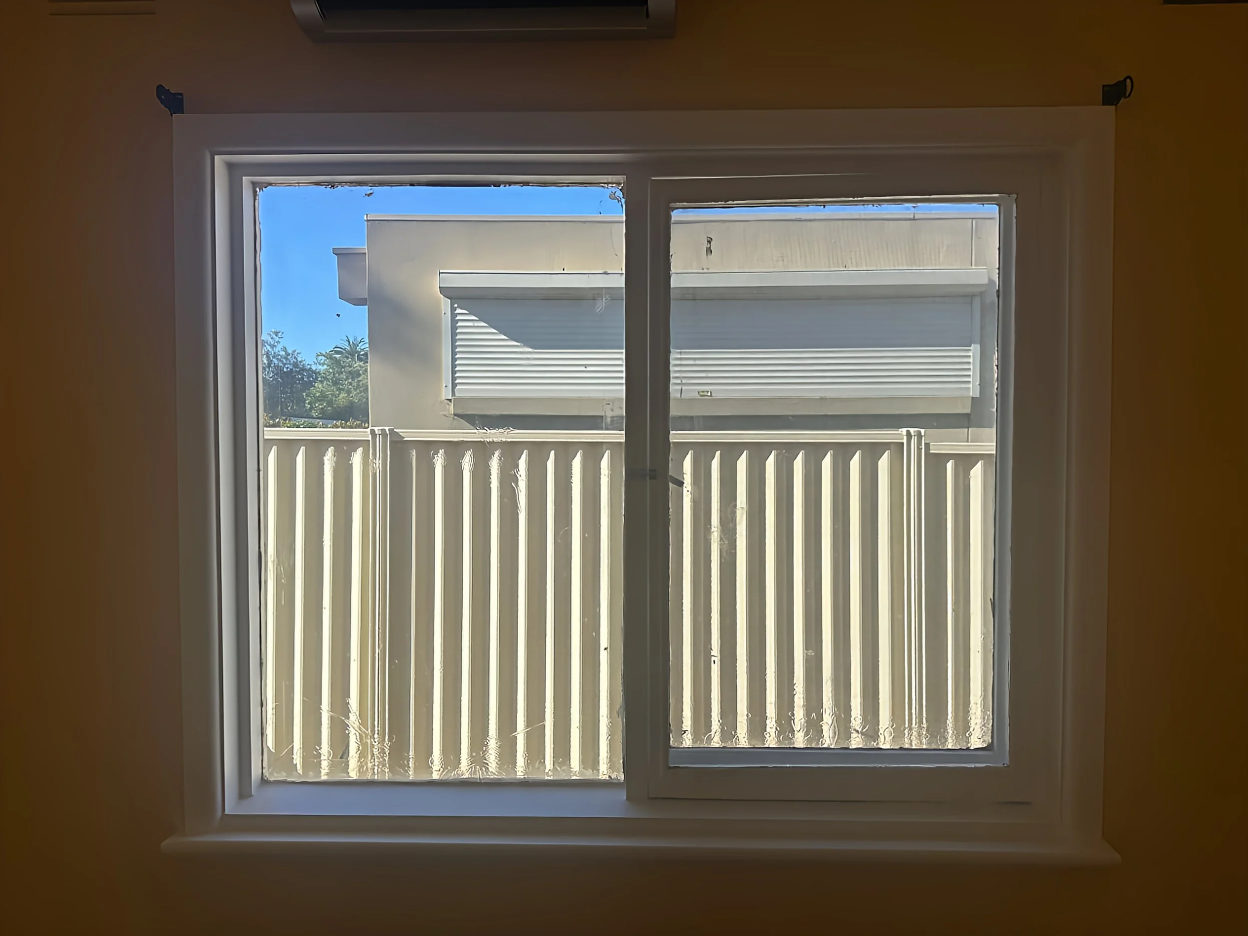 View through a window showing a white fence outside with a neighboring building and blue sky.