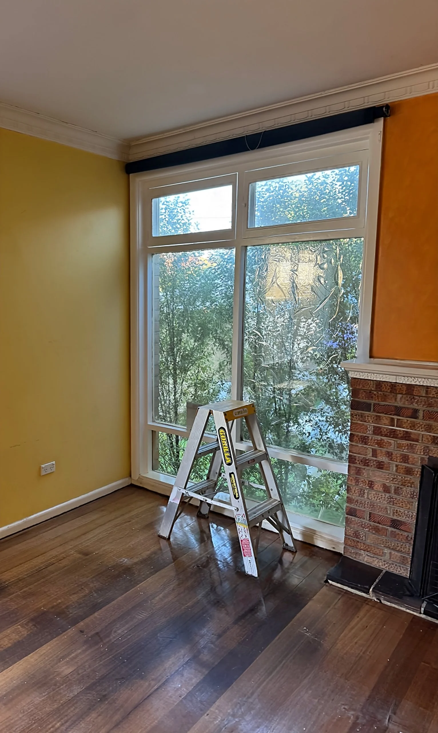 Living room with large window overlooking trees, yellow wall, brick fireplace, and wooden floor. A small step ladder is positioned near the window.