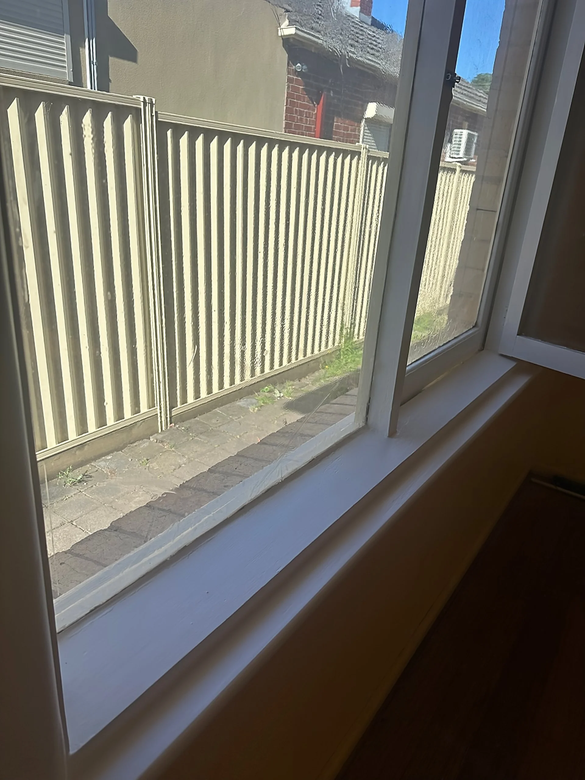 View through a closed window showing a beige fence, brick pathway, and neighboring house with red brick wall and outside air conditioning unit.