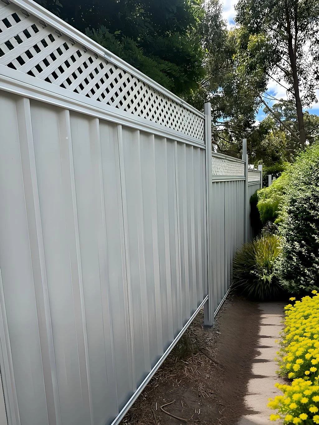 A white metal fence with a lattice top, running alongside a garden with yellow flowers and green shrubs, under a partly cloudy sky.