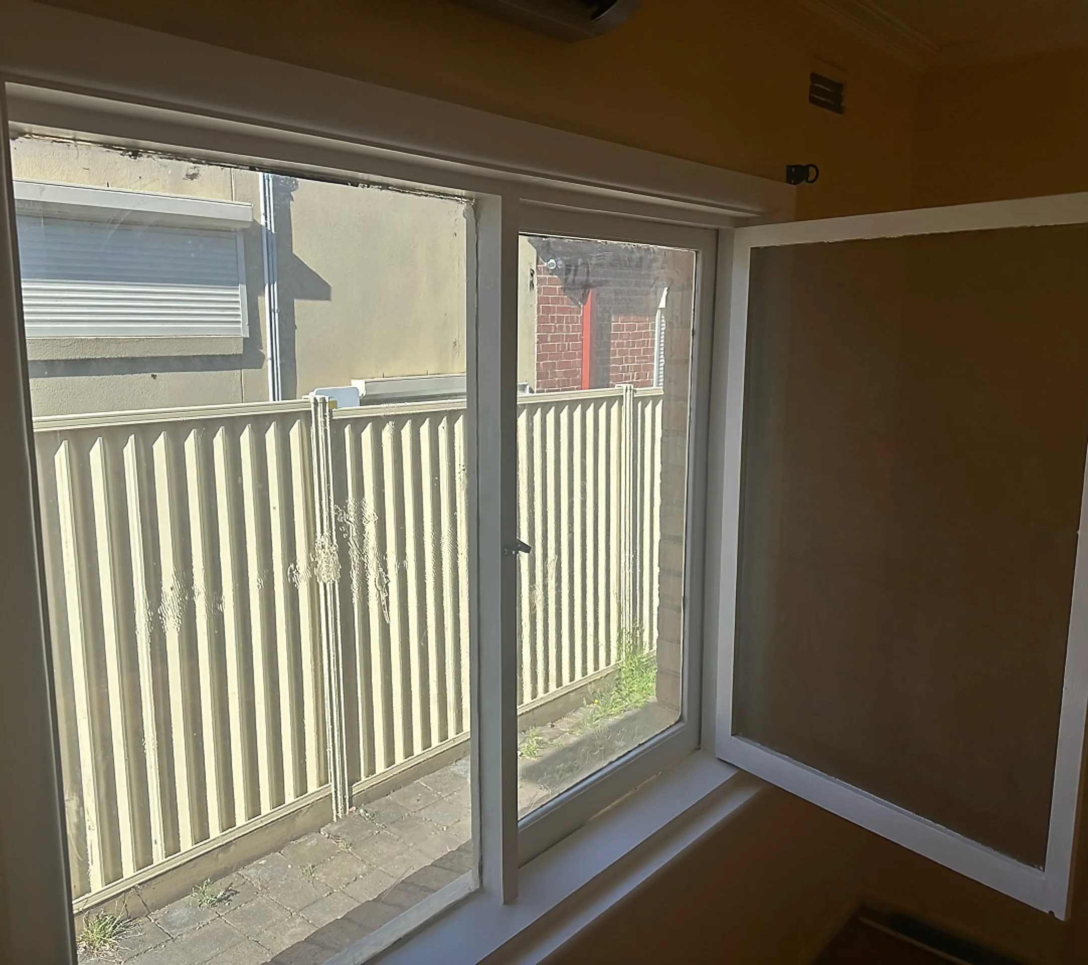 View through a large glass window showing a beige wooden fence, pavement, and the side of neighboring buildings with a window and brick wall.