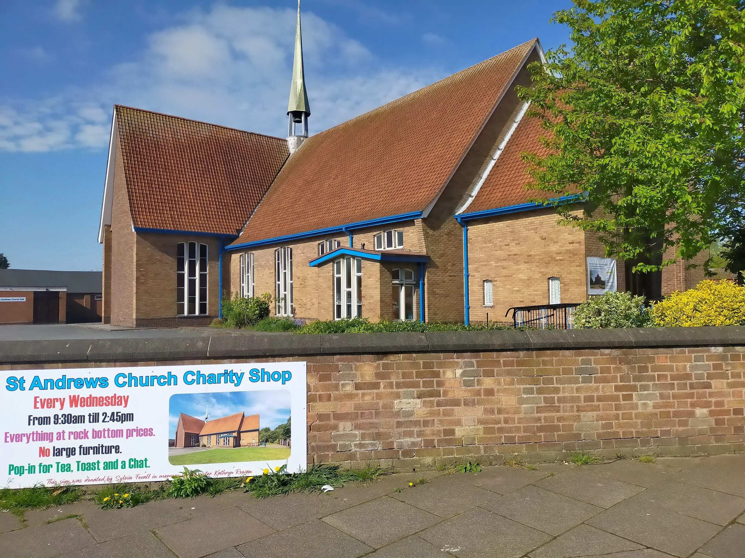 A brick church with a tall steeple under a partly cloudy sky, surrounded by greenery, with a promotional sign for a charity shop in front.