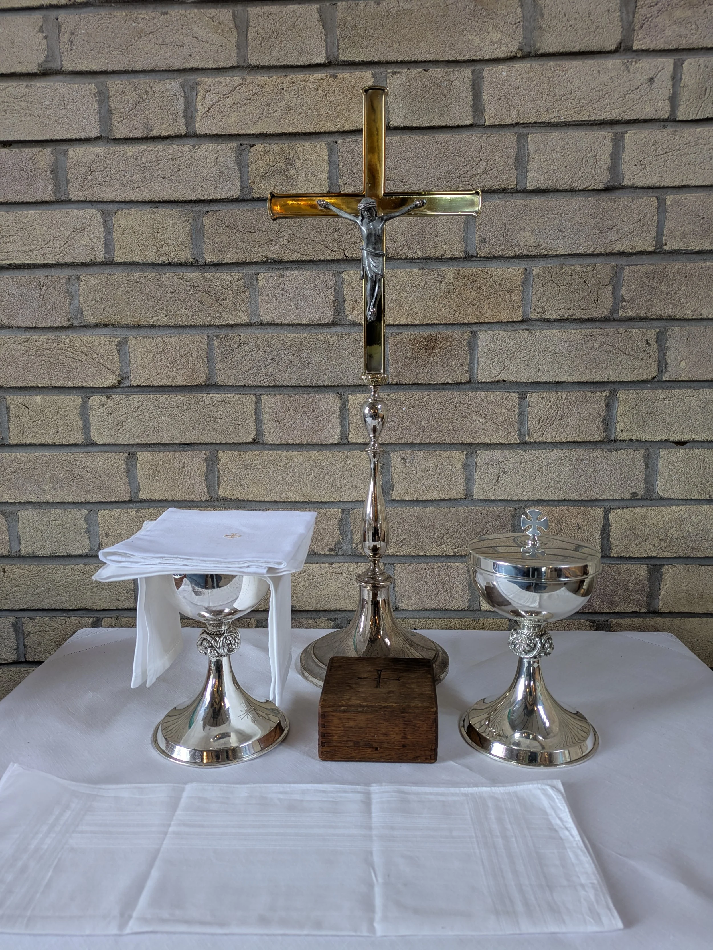 Christian altar with a crucifix, a wooden box, a silver chalice, and a ciborium on a white cloth against a brick wall.