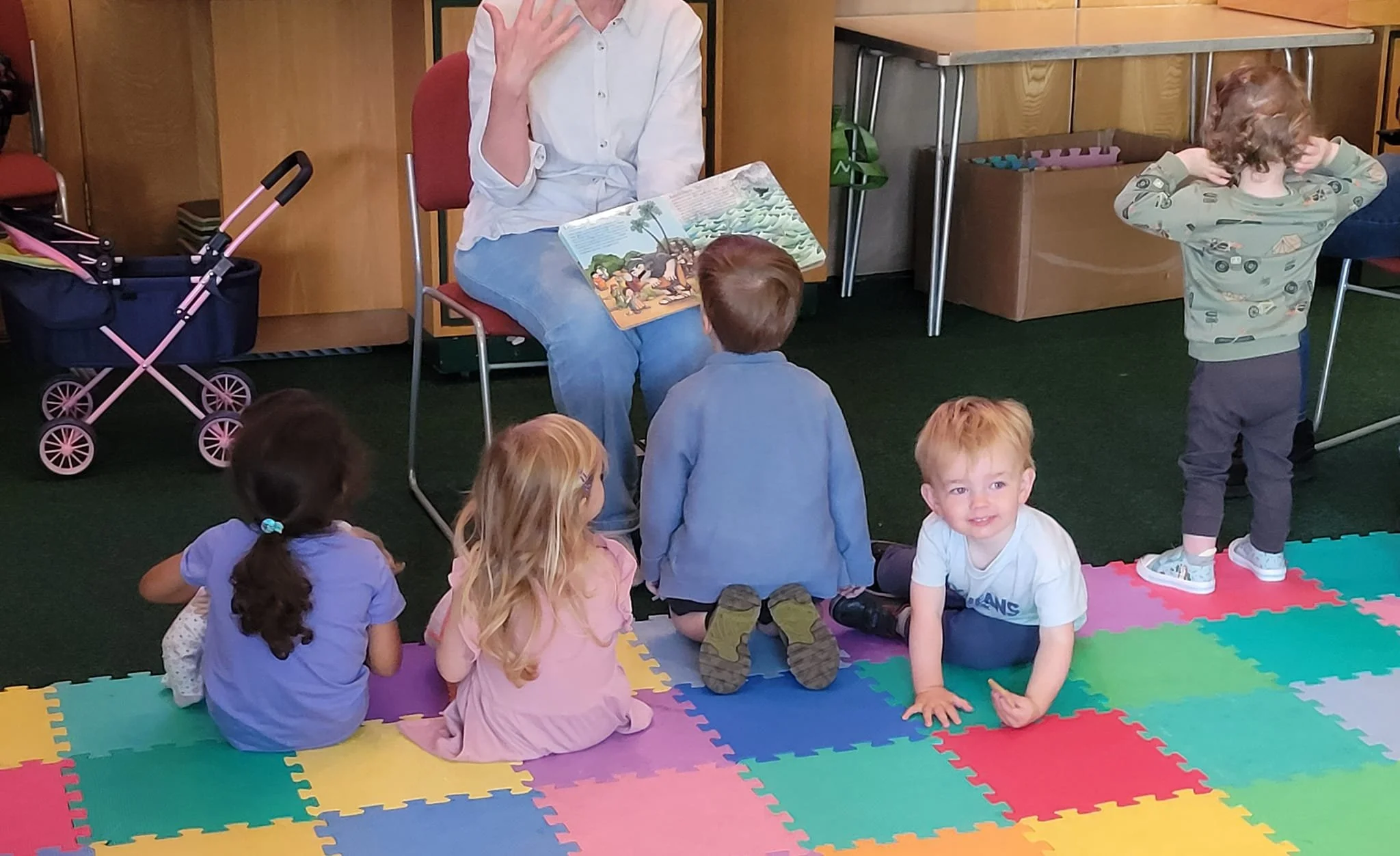 A group of young children sitting and kneeling on a colorful foam mat, listening to a woman reading a picture book in a community group.