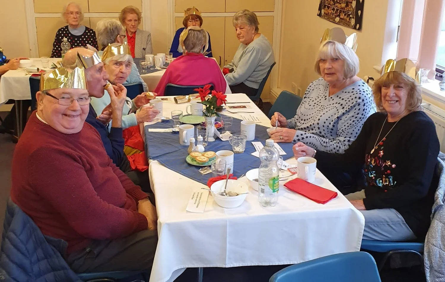 A group of elderly people at a holiday party, some wearing party hats, sitting around a table with food, drinks, and Christmas decorations.