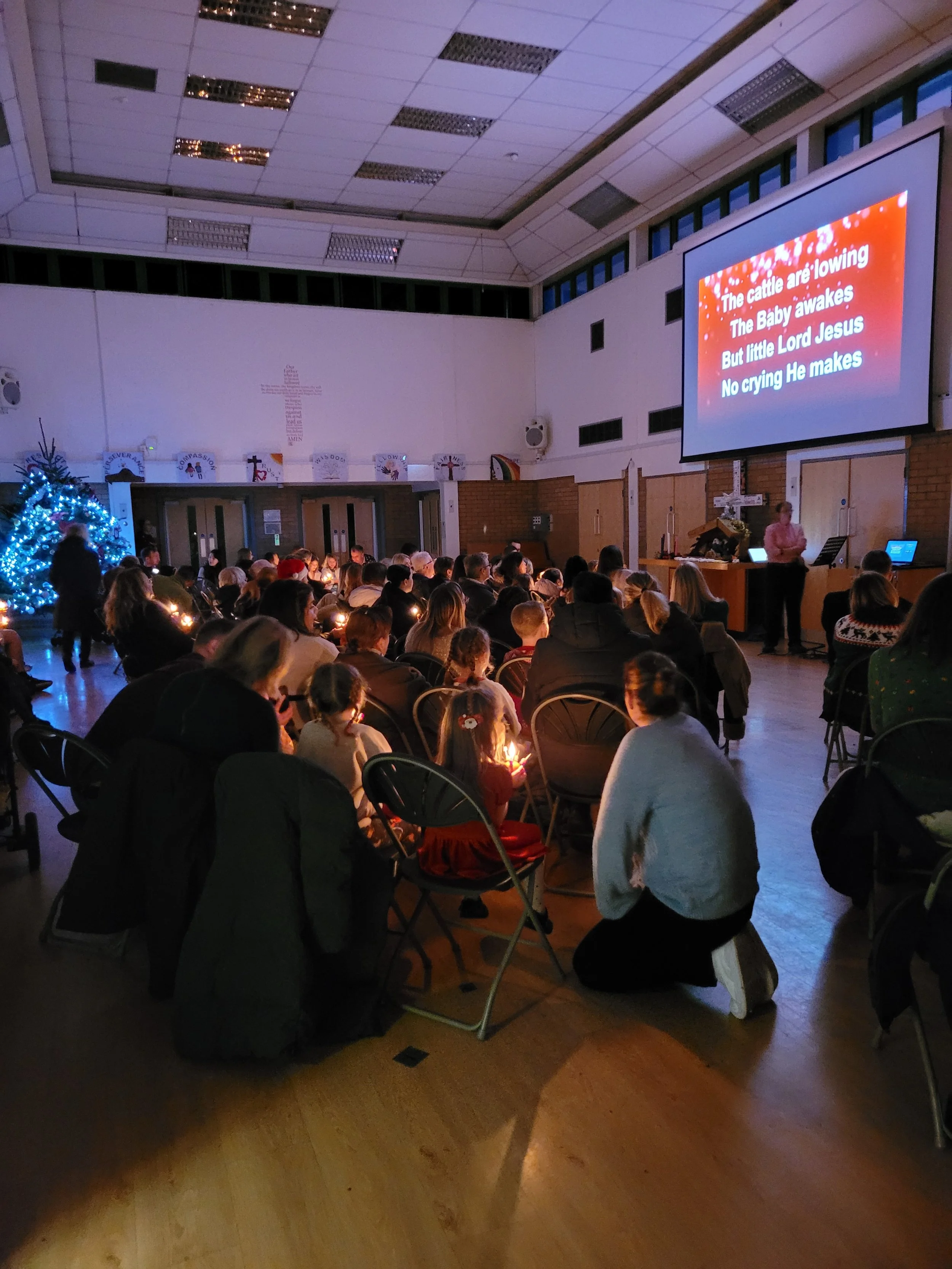 A church hall filled with people sitting in chairs, many holding candles, during a Christmas service. A Christmas tree with lights is visible on the left. On the large screen at the front, Christmas song lyrics are displayed, and a person is standing near a keyboard.