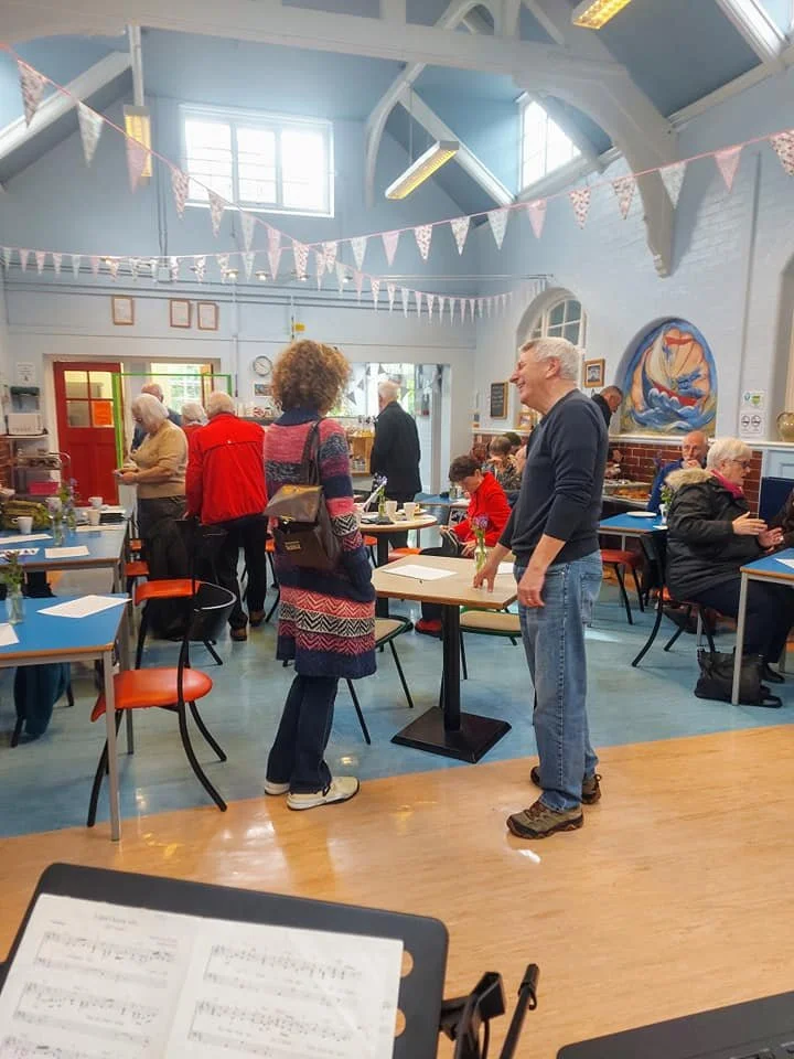 People in a bright, spacious room with high ceilings, decorated with pink and white bunting, engaging in conversations and enjoying drinks on tables.