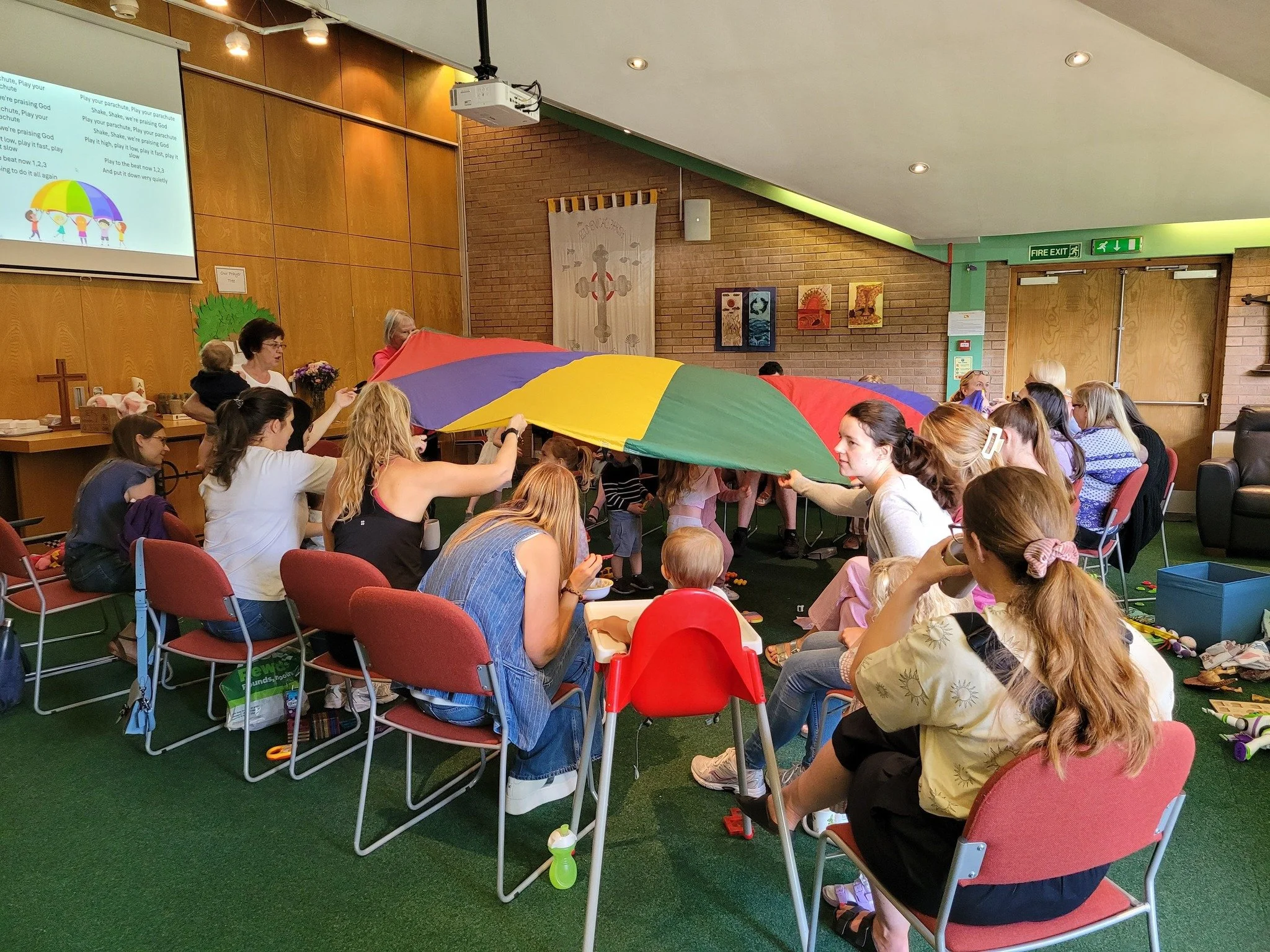 Children and adults gathered indoors, holding a colorful parachute during a group activity, with a screen displaying text and some artwork on the walls in the background.