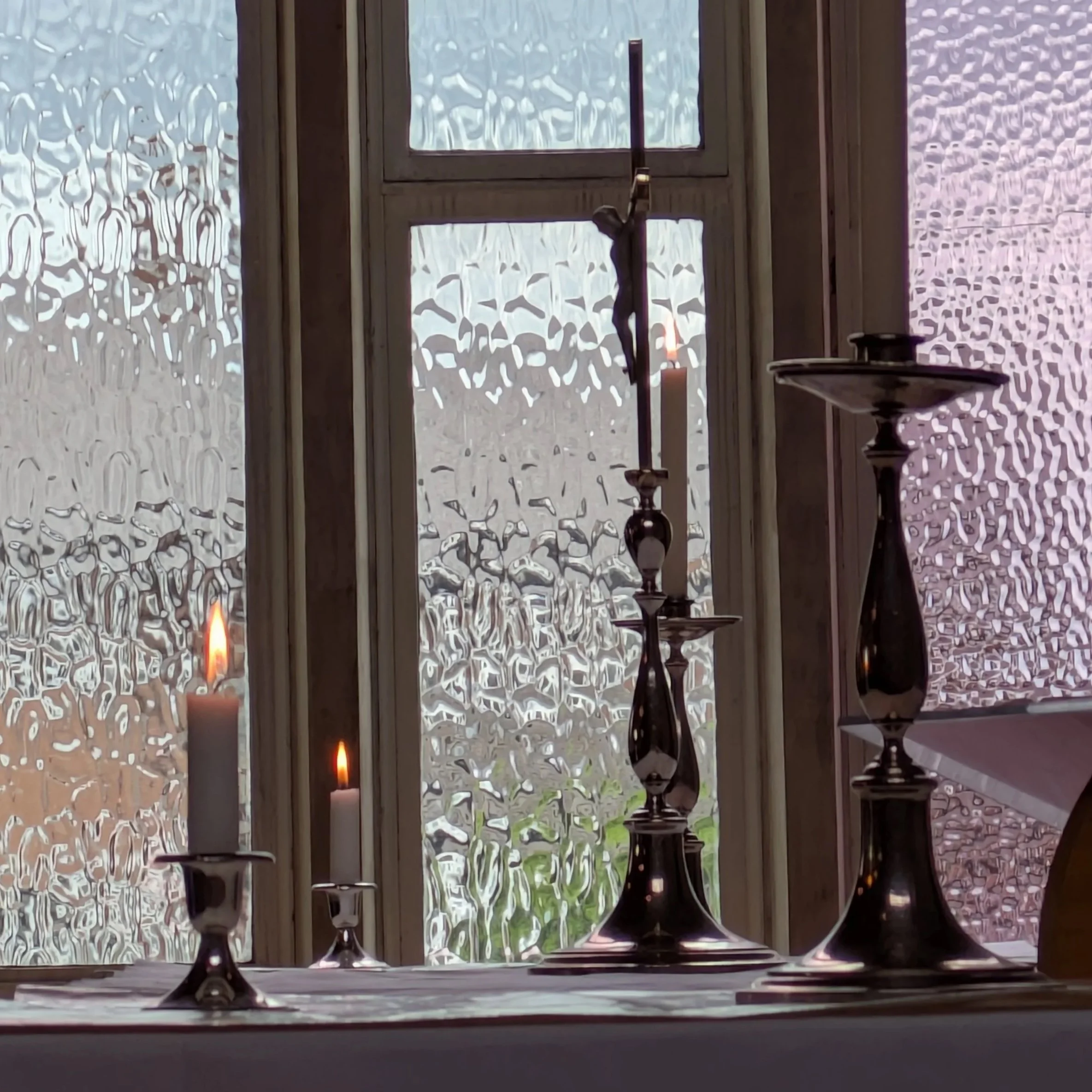 Religious altar with lit candles, crucifix, and decorative candlesticks in front of frosted window.