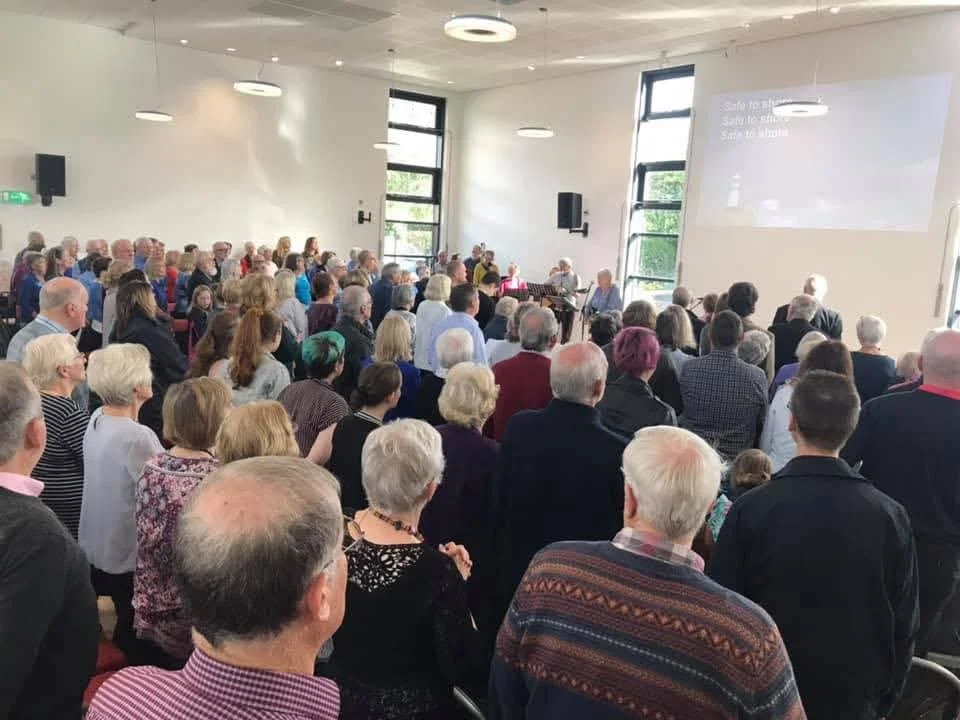 A large gathering of people in a spacious, well-lit room with white walls, windows, and ceiling lights, attending an event or presentation.