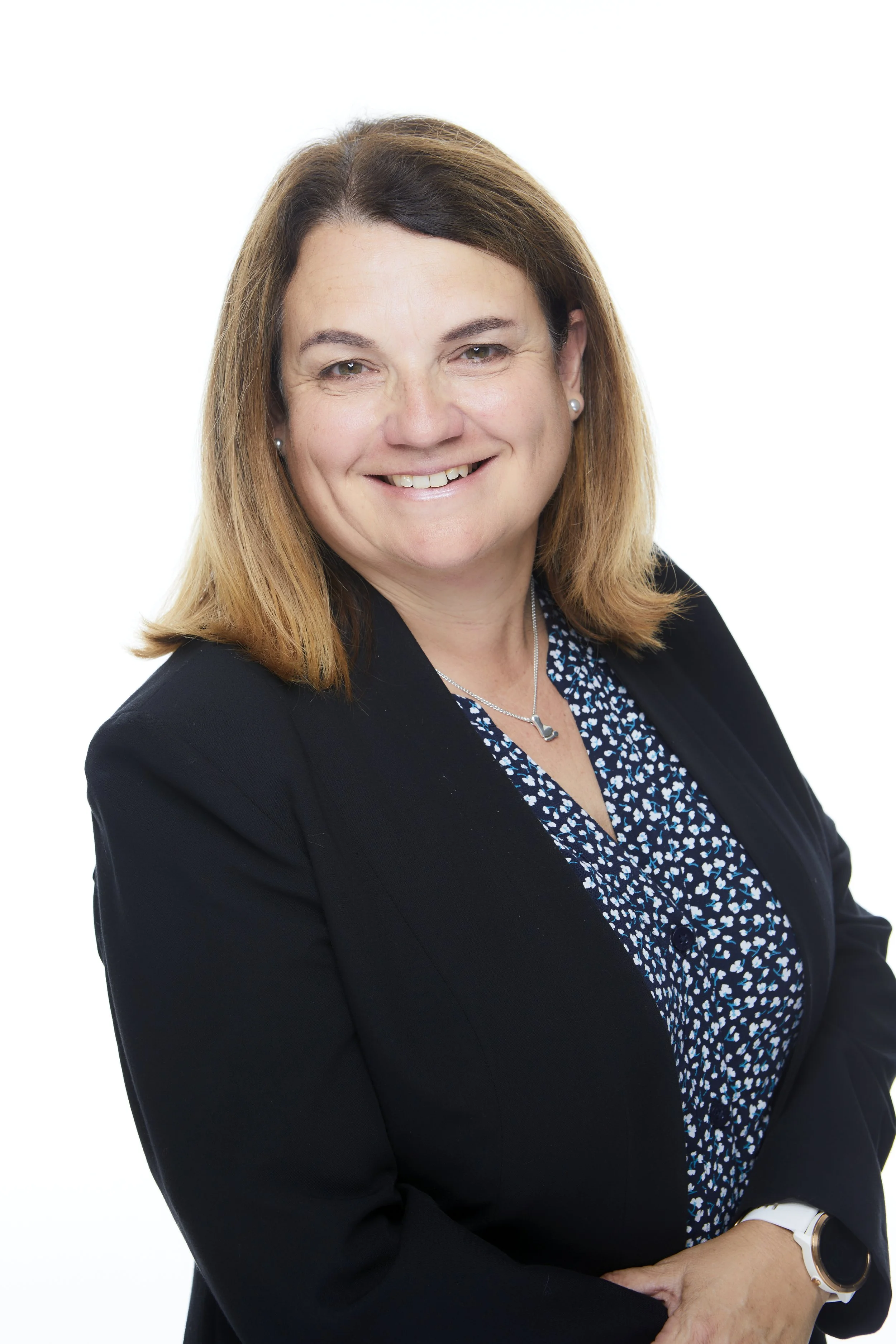 A woman with shoulder-length blonde hair smiling, wearing a black blazer and a black and white patterned blouse, against a plain white background.