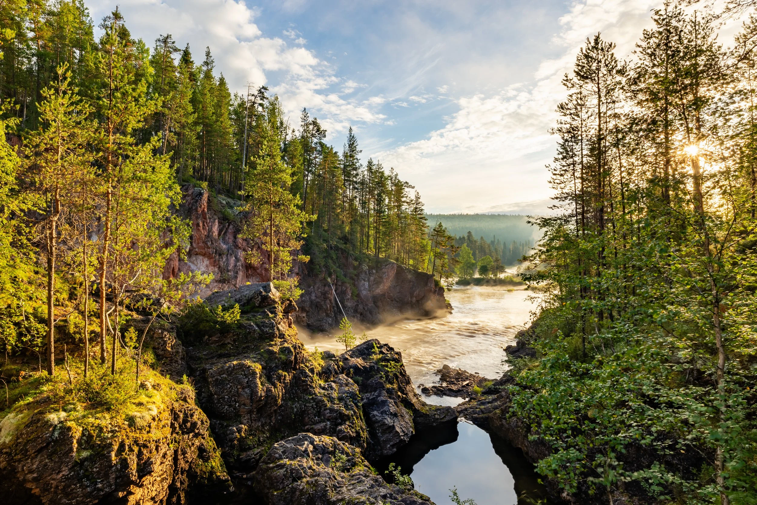 Foresta con alberi verdi, rocce, un fiume con vapori e il cielo con nuvole
